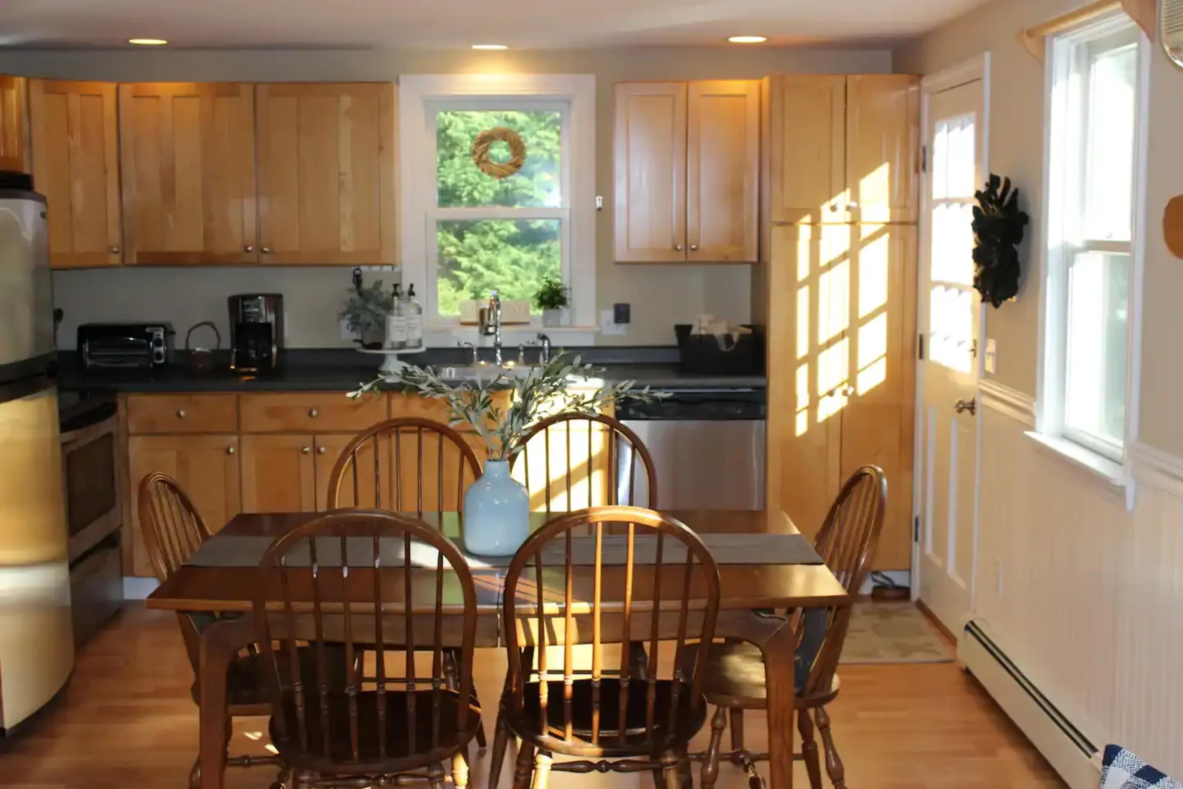 A dining area with a wooden table and chairs is positioned in front of a kitchen featuring light wood cabinetry and a window with natural light streaming in.