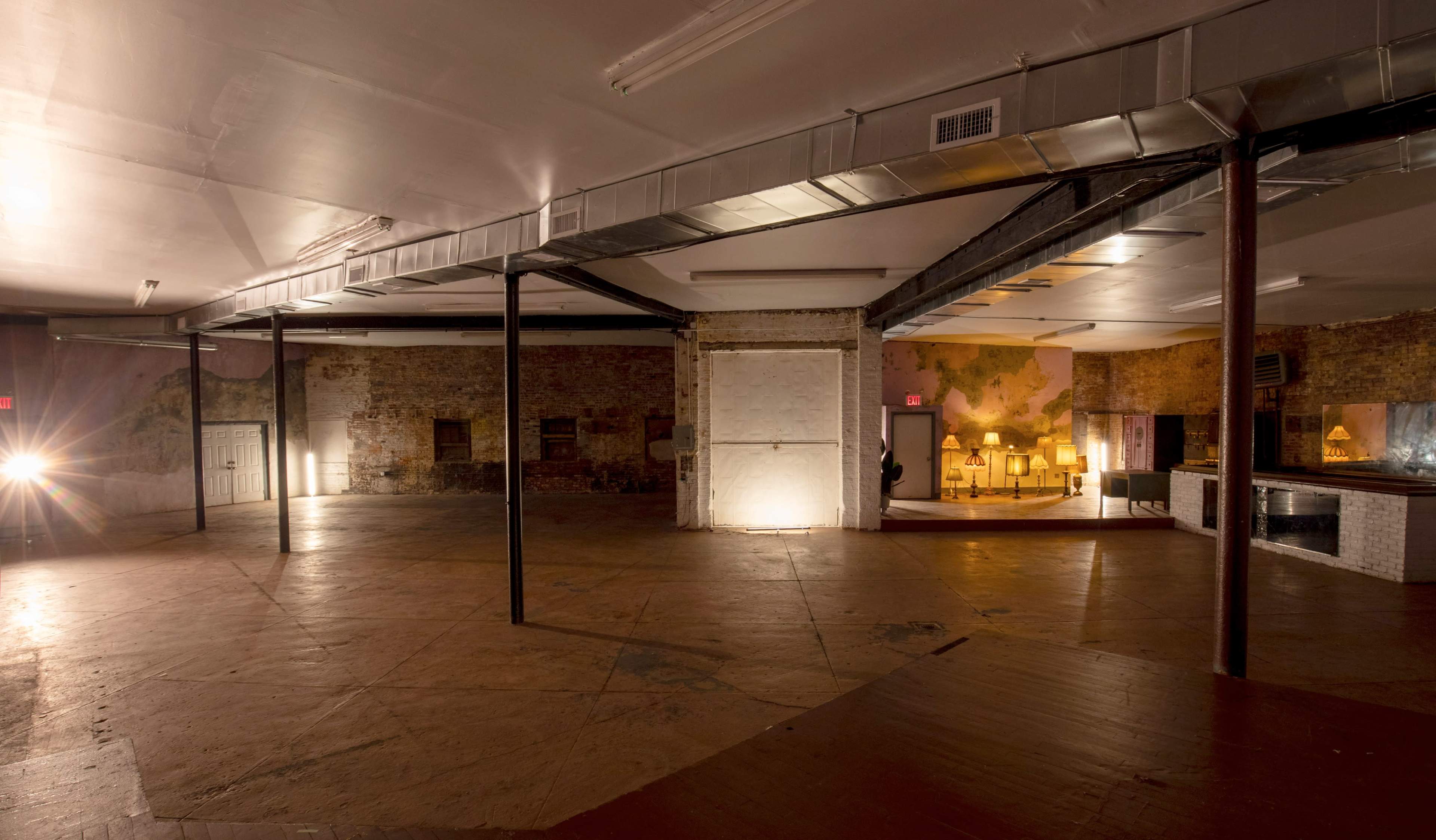 The image shows an empty industrial-style room with exposed brick walls, wooden floors, and various light sources illuminating different areas.