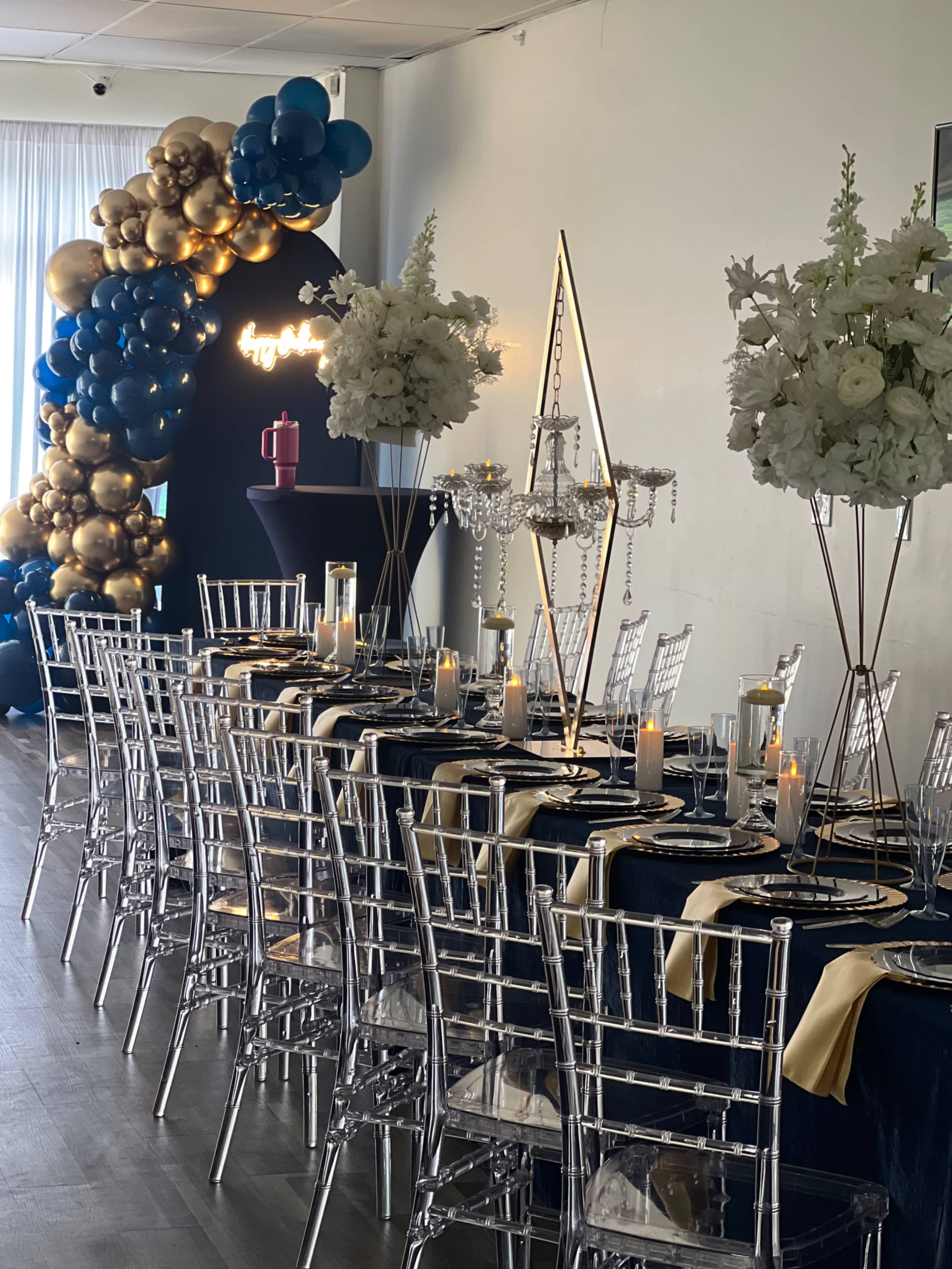 A long banquet table is elegantly set with black and gold decor, surrounded by clear acrylic chairs and adorned with flowers and candles under a balloon arch in the background.