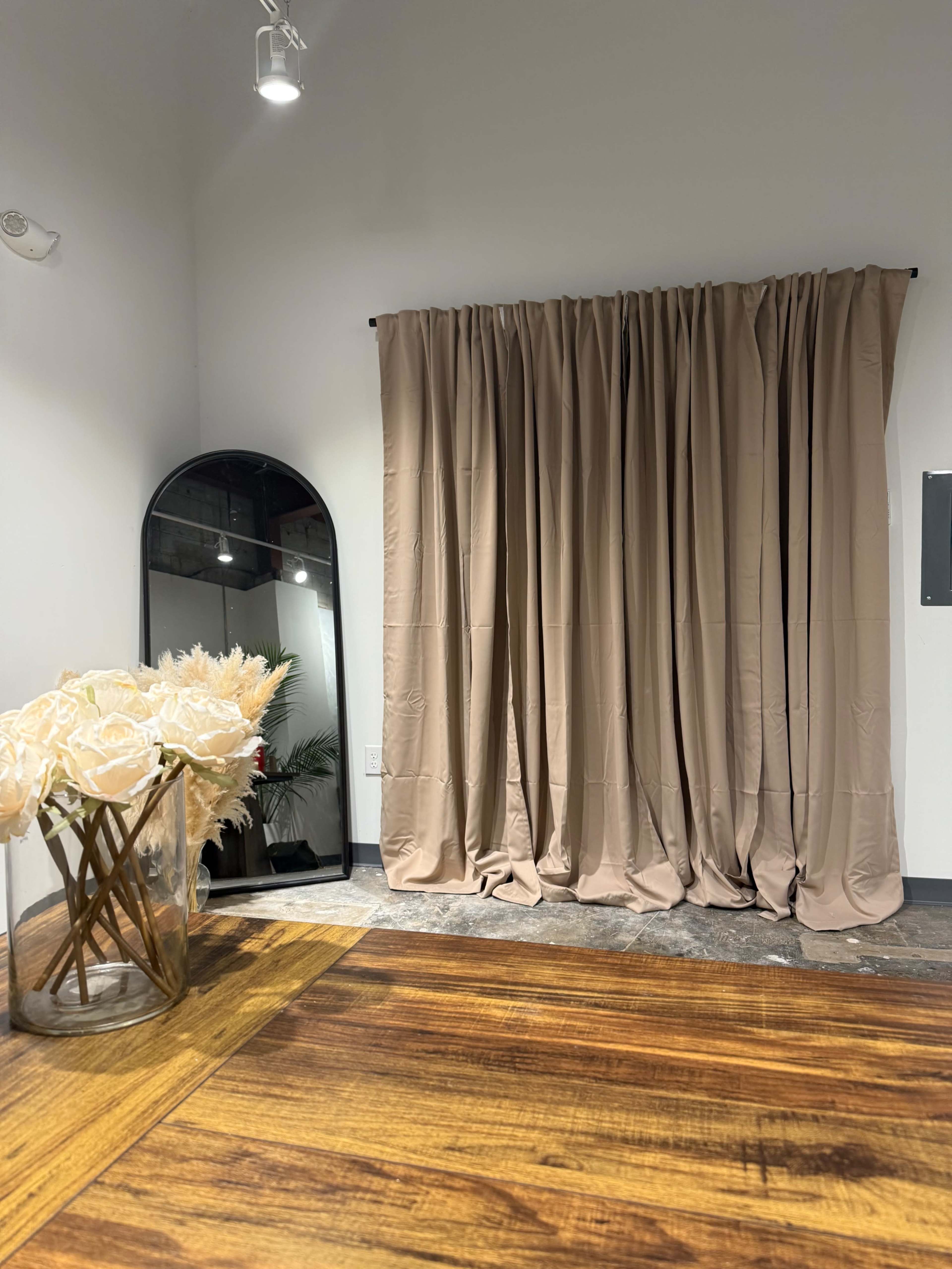 The image shows a room with a wooden floor, a large mirror, a beige curtain draped against the wall, and a vase of flowers on a table.