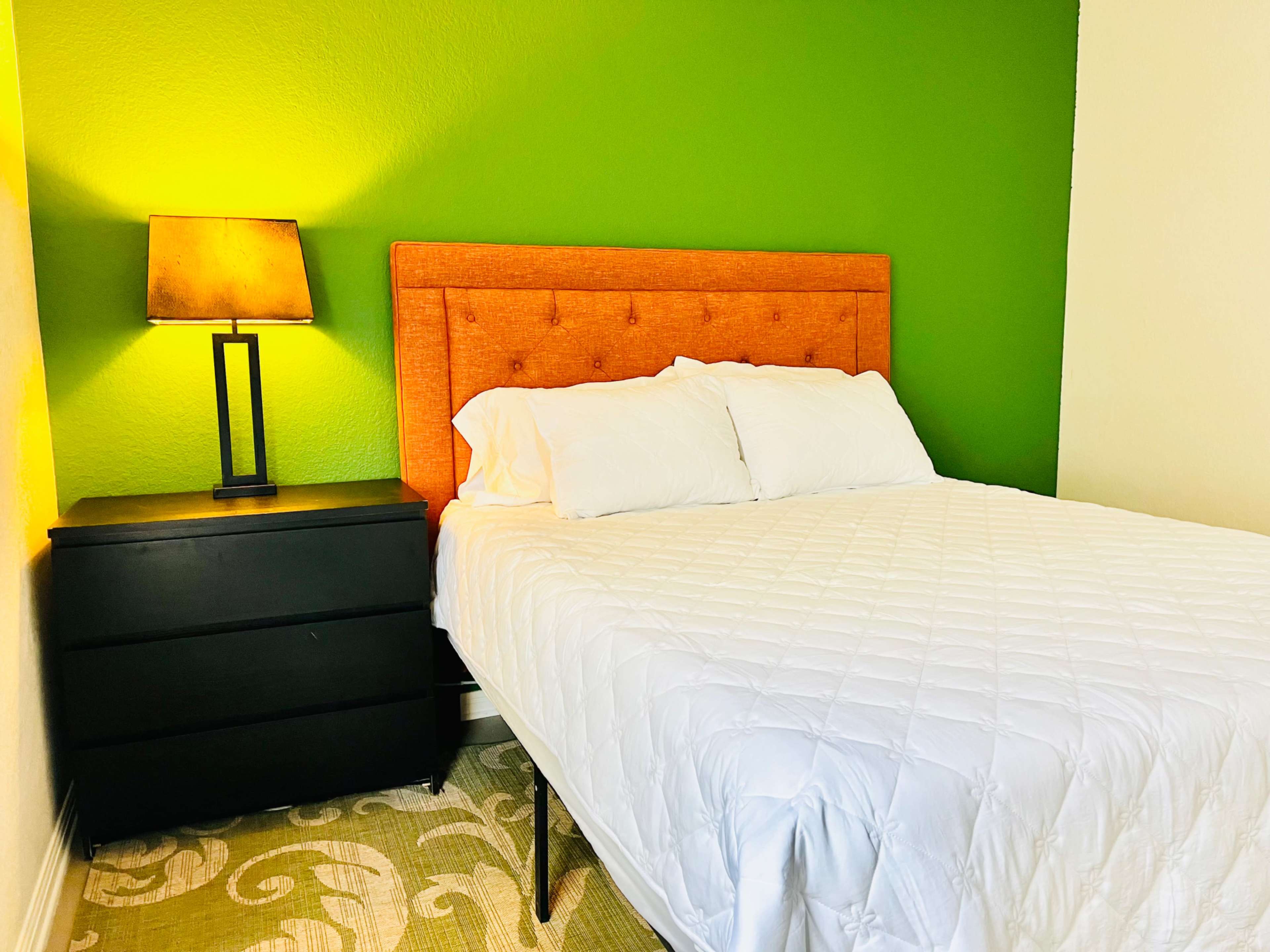 The image shows a bedroom with a green accent wall, an orange upholstered headboard, a white bedspread, a black nightstand, and a lamp beside the bed.