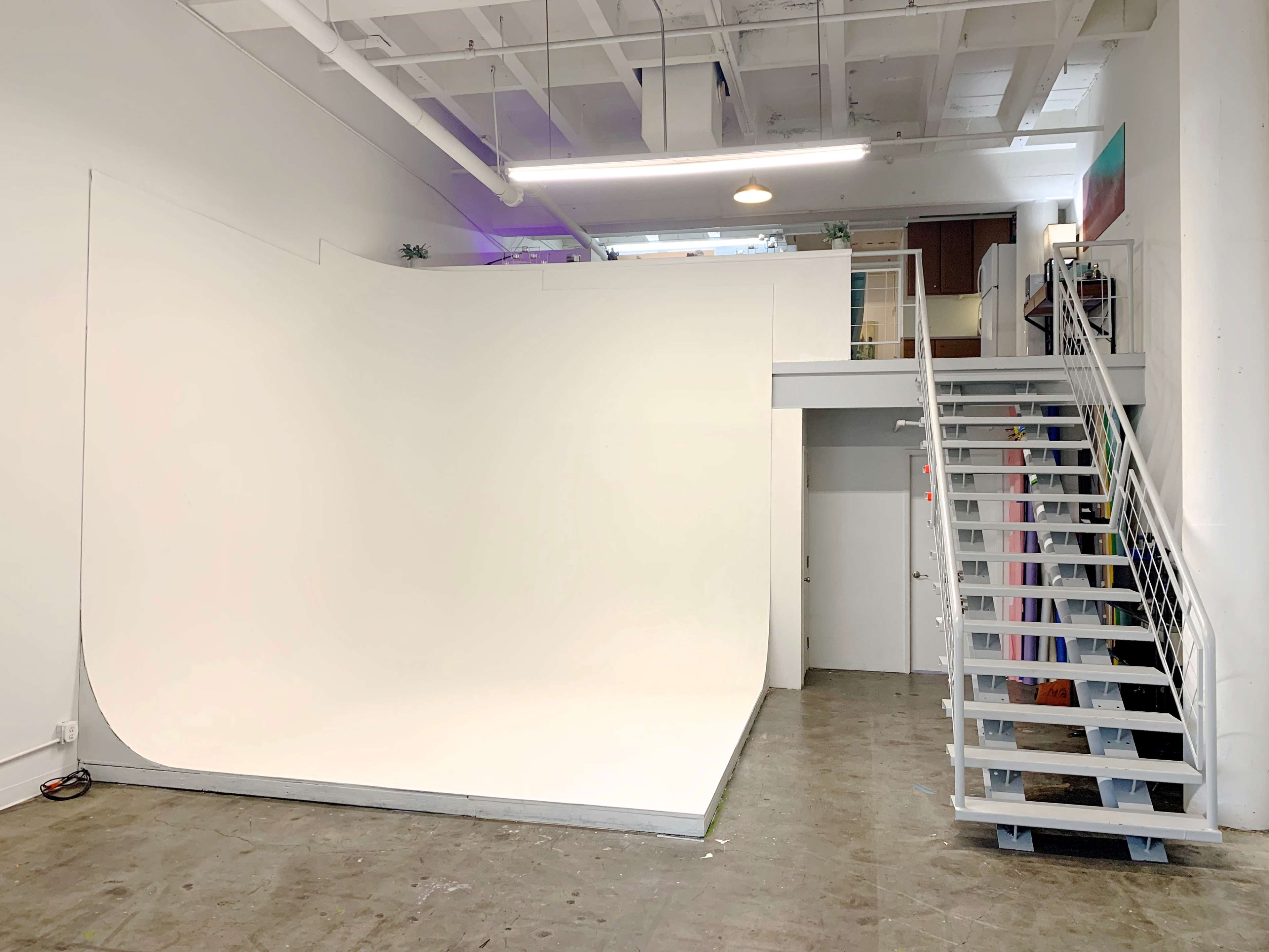 A bright, minimalist studio space with a large white background and a staircase leading to an upper level.