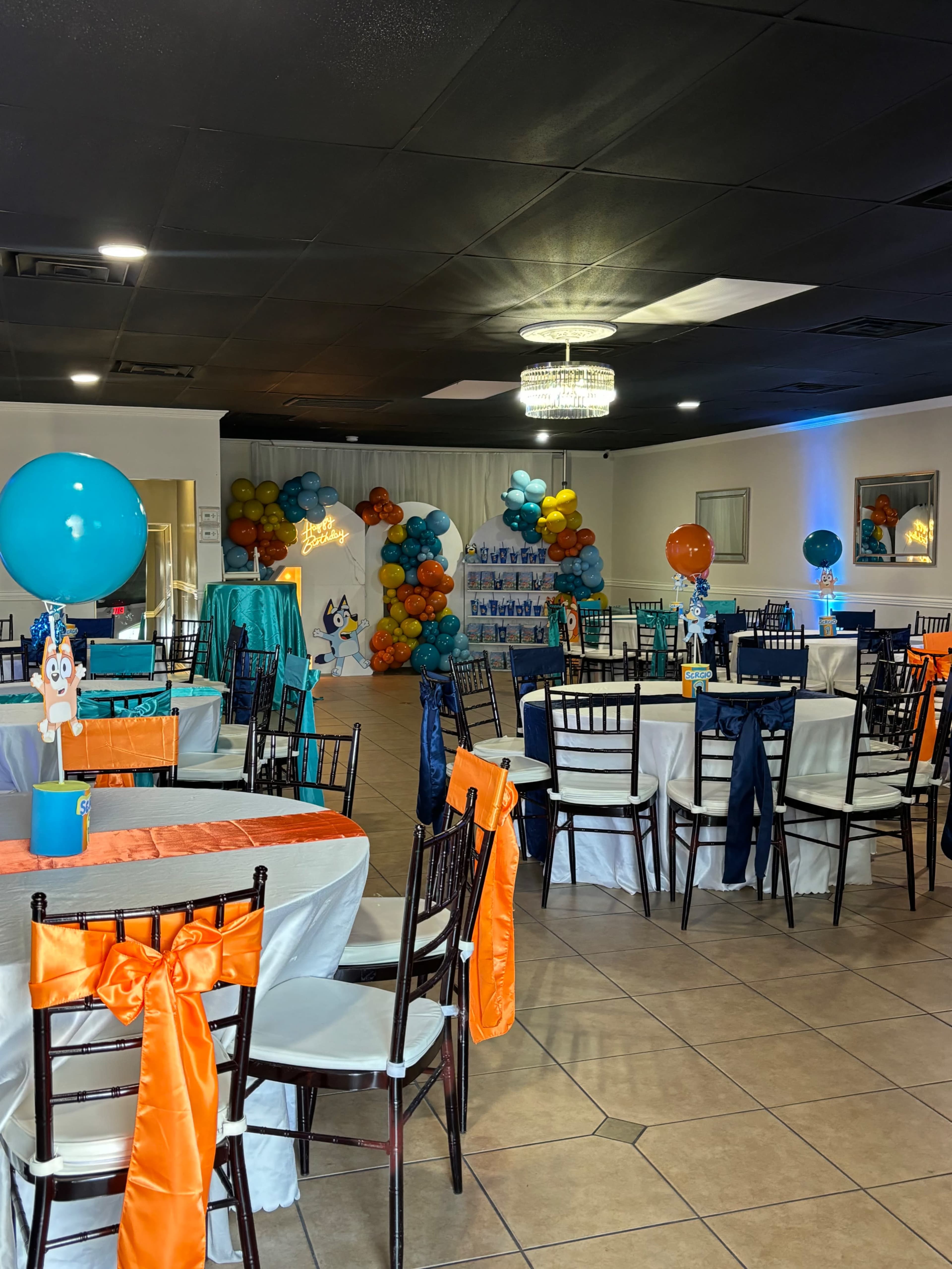 The image shows a decorated event space with tables set for a celebration, featuring orange and blue themes with balloons and centerpieces.