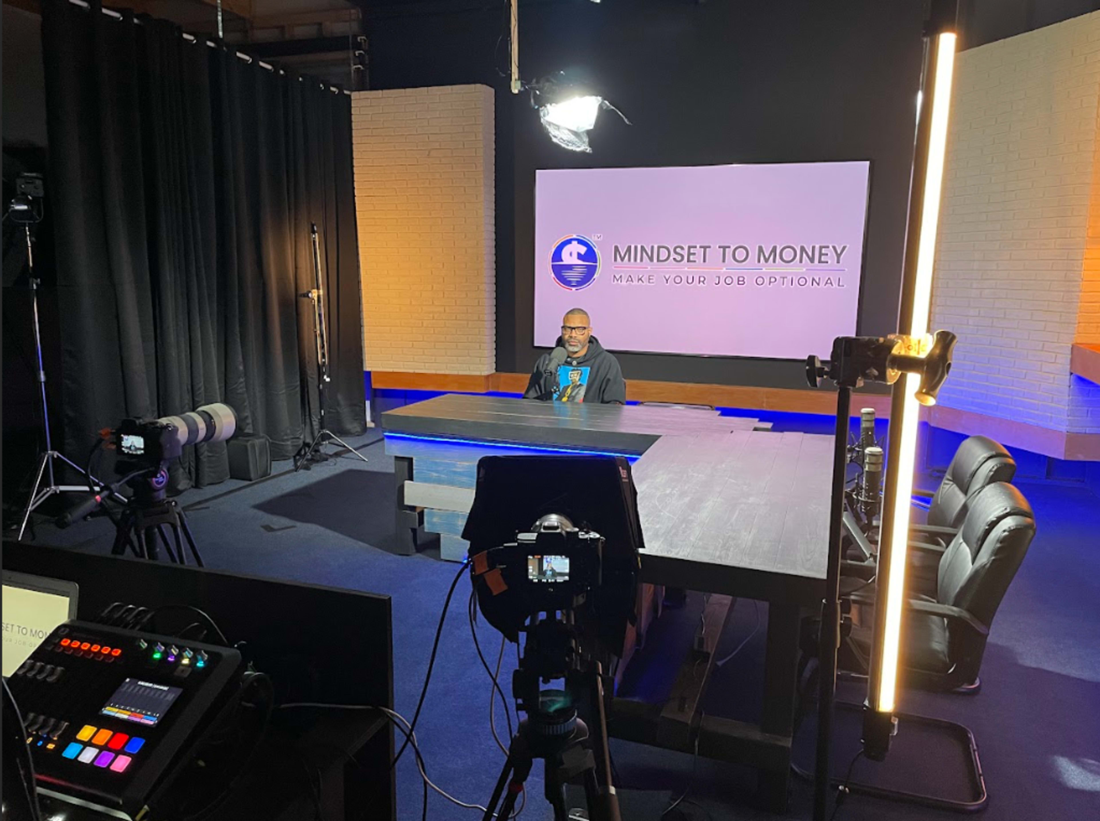 A speaker sits at a large table in a well-lit studio, with cameras and equipment set up for a production labeled "Mindset to Money."