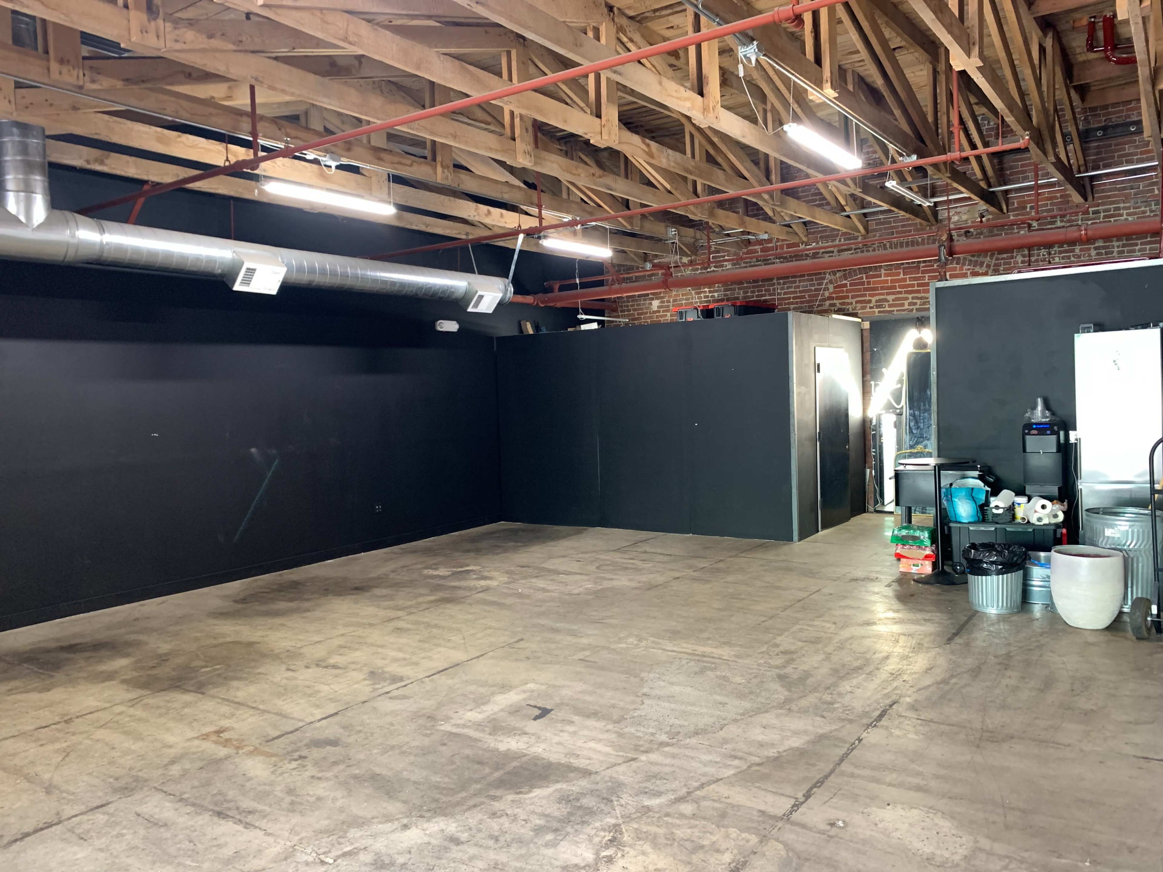 An empty industrial space with exposed wooden beams, a concrete floor, and black walls, featuring a faint outline of a partition and various equipment in the corner.