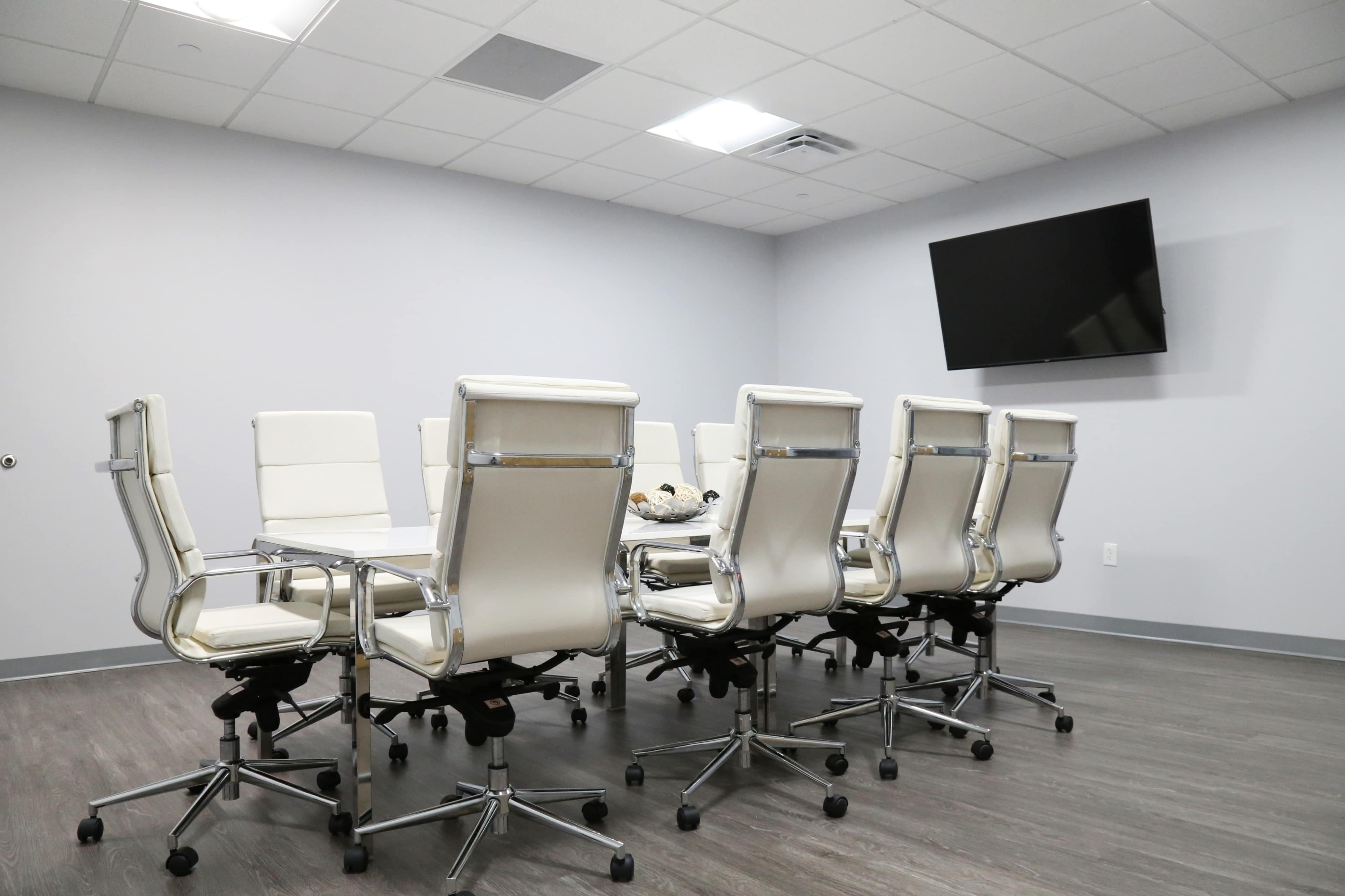 A modern conference room features a long table surrounded by eight white leather office chairs and a wall-mounted television.