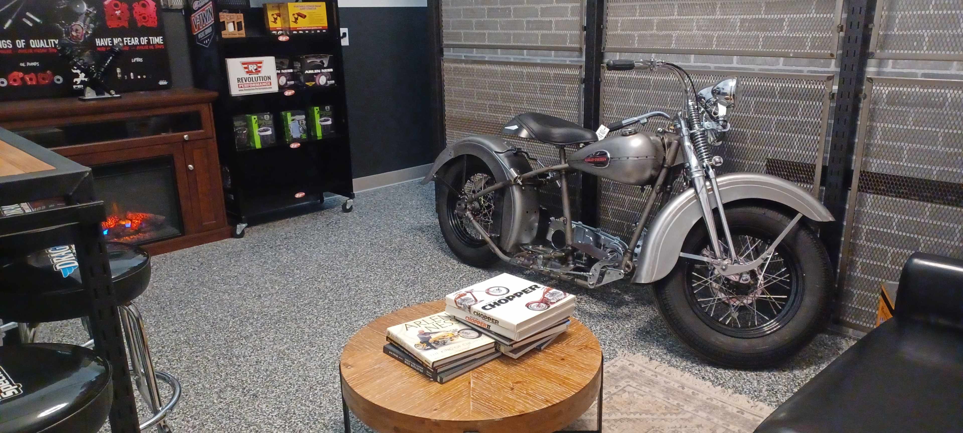 A vintage motorcycle beside a round wooden coffee table stacked with magazines in a modern garage-like space.