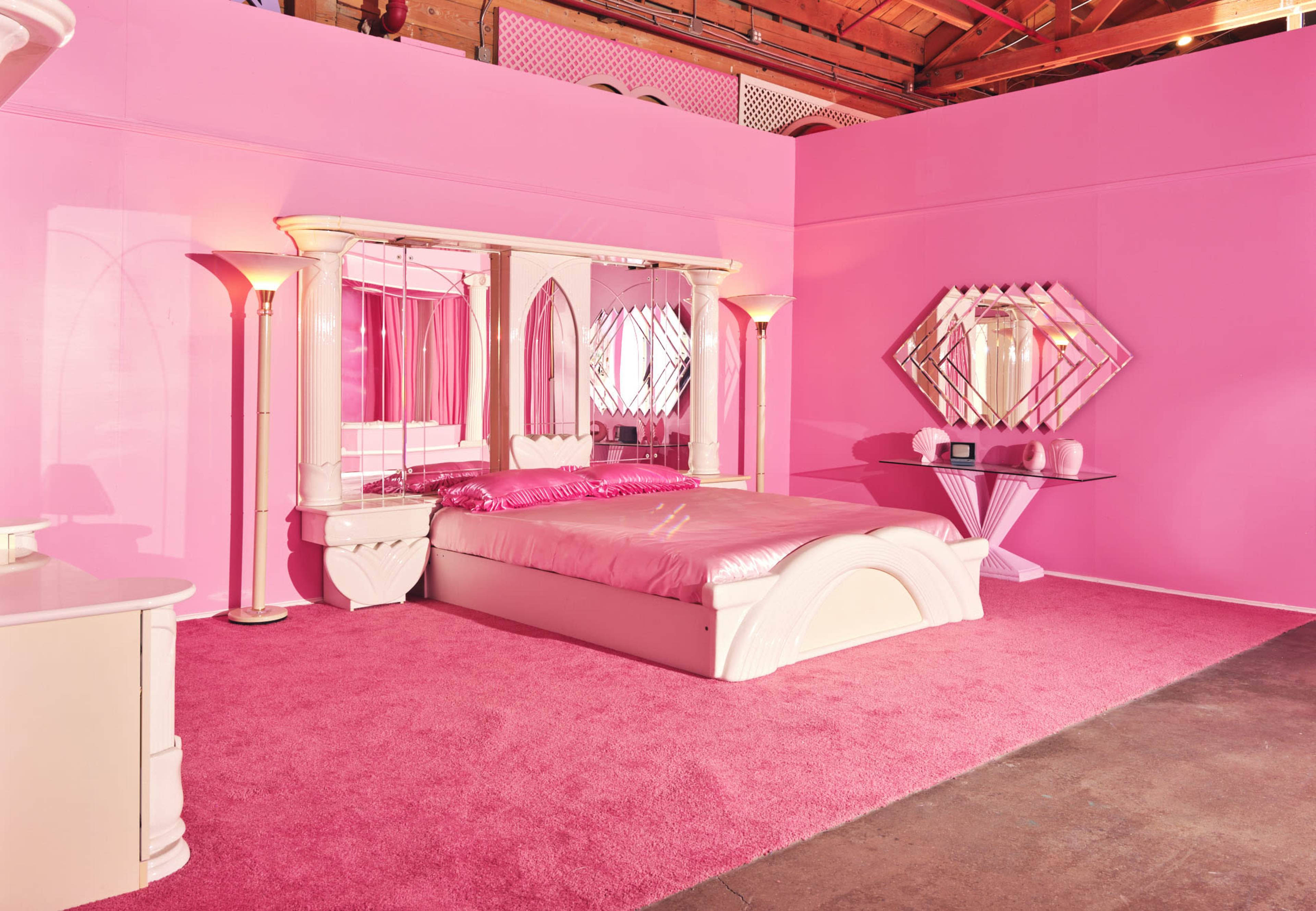 The image shows a pink-themed bedroom featuring a bed with a pink comforter, matching walls, and mirrored furniture.