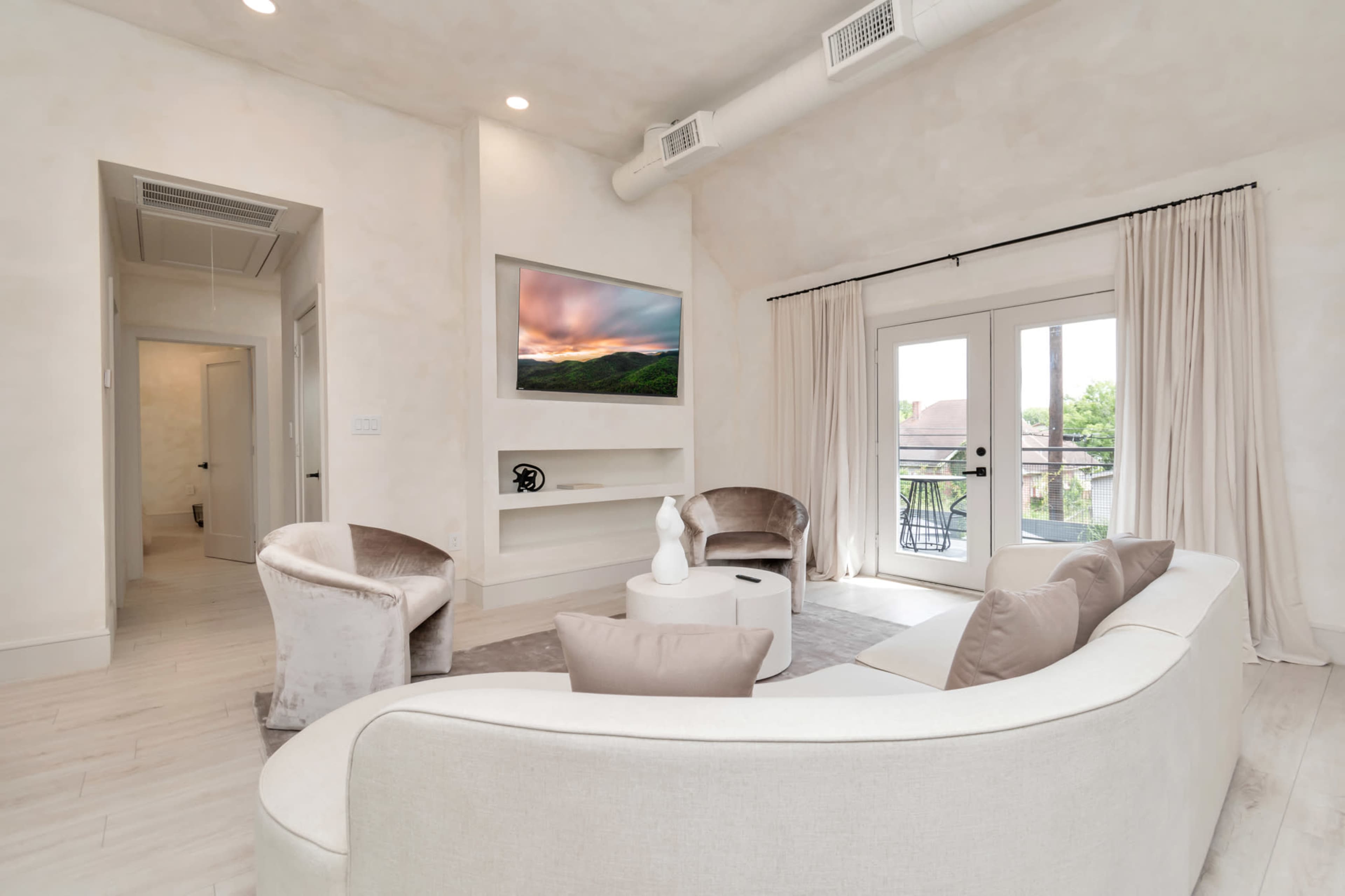 The living room features a curved sofa, two armchairs, a coffee table, and a television mounted on the wall, with large windows draped in curtains.