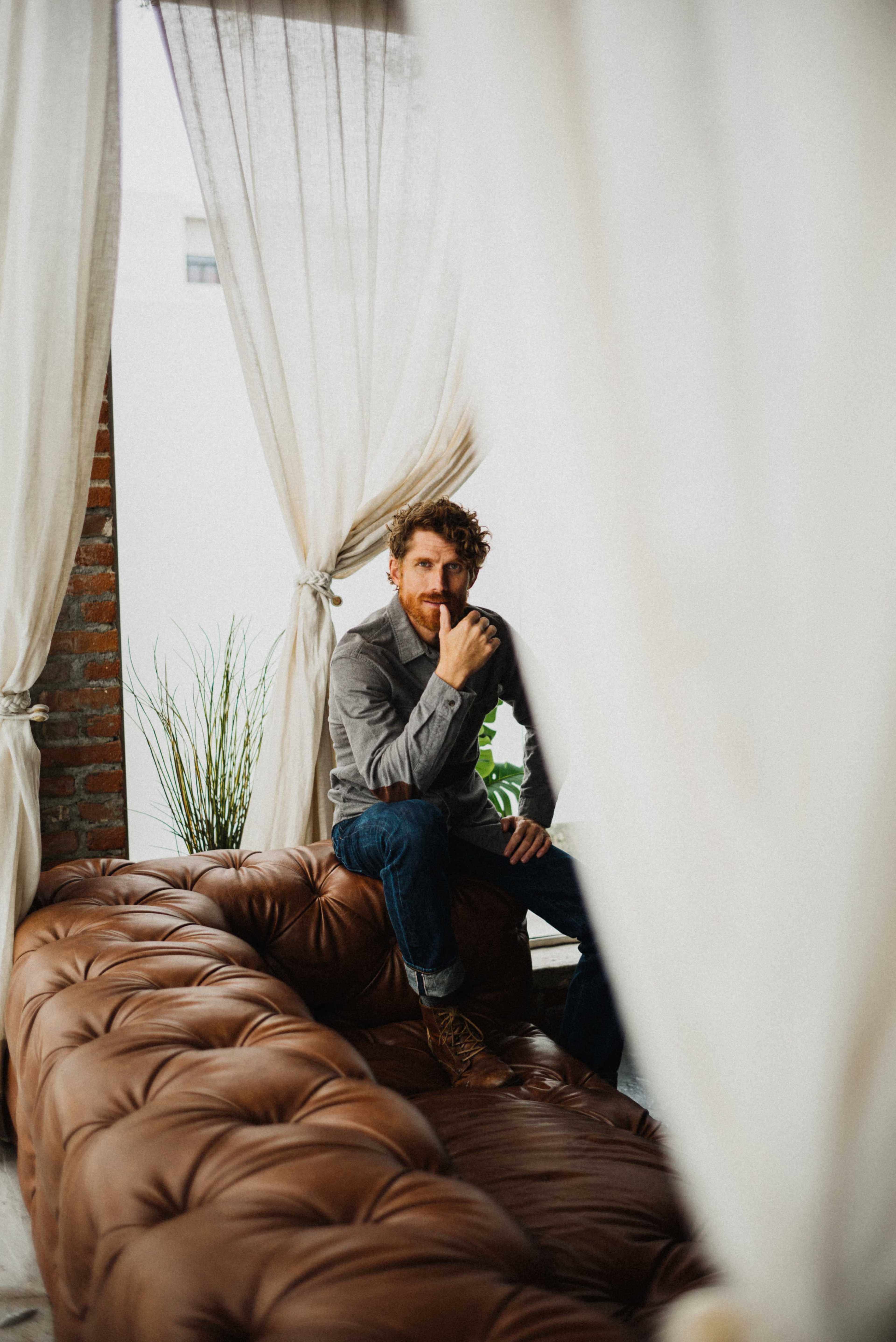 A man sits thoughtfully on a brown leather couch, framed by white curtains and plants in the background.