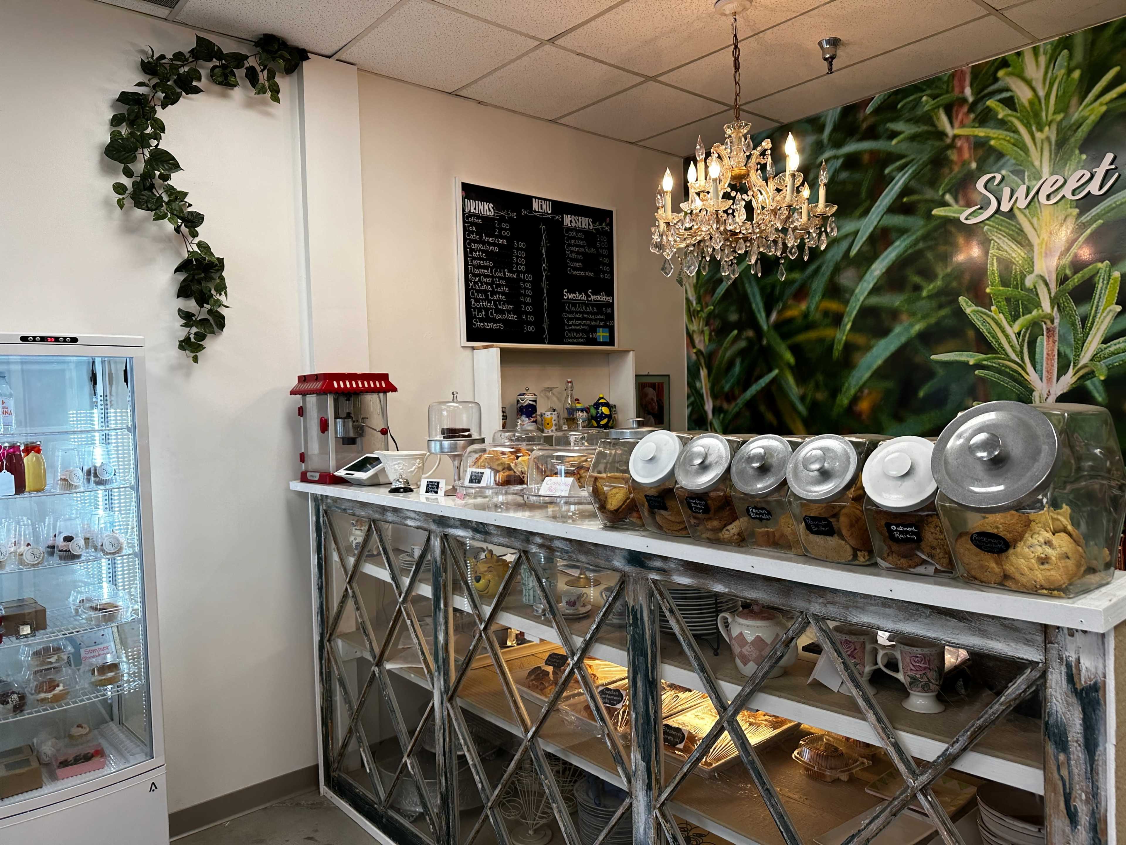 A bakery counter displays a variety of baked goods in clear containers, with a menu board and a chandelier overhead.