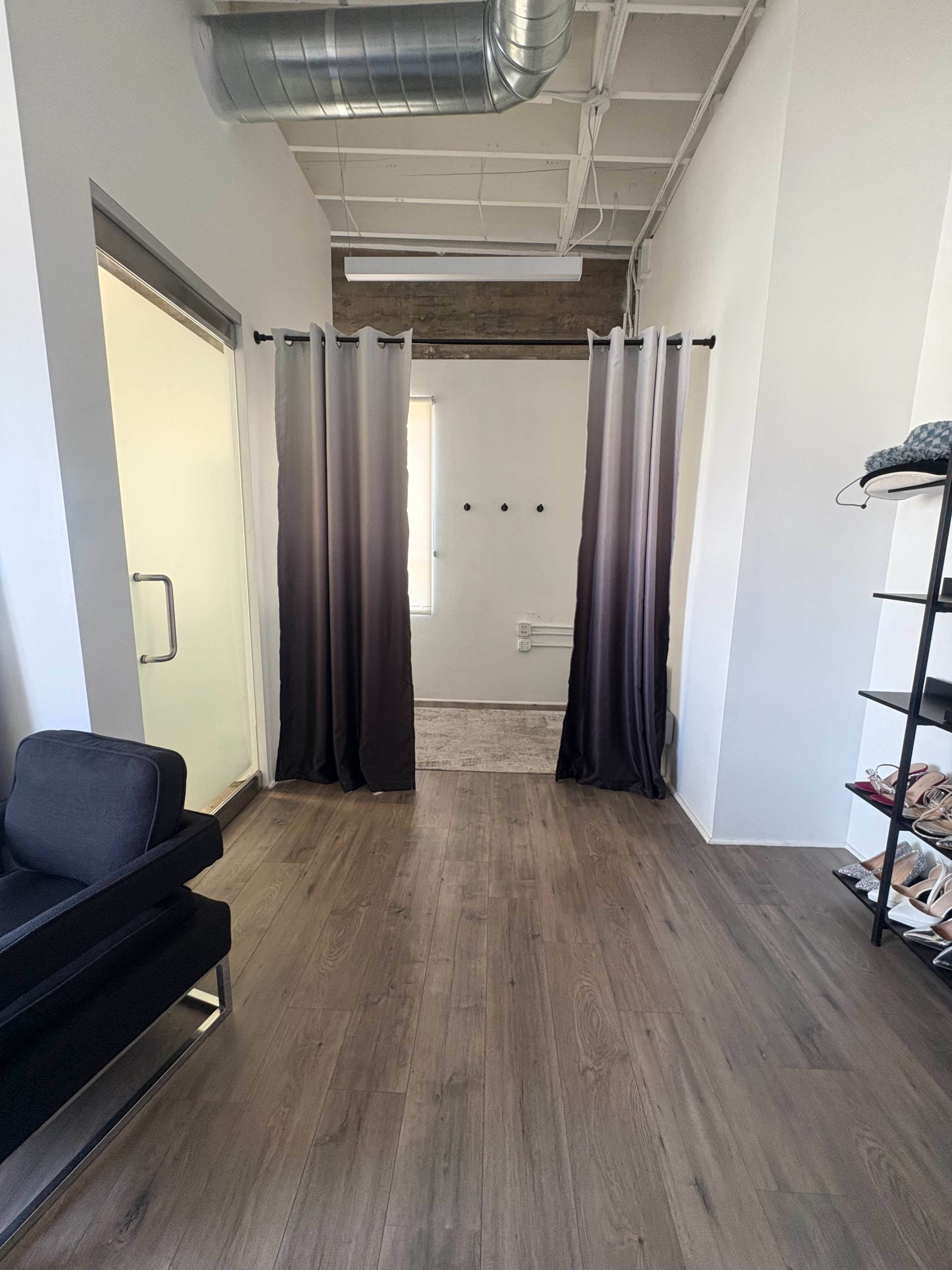 The image shows a spacious room with gray curtains separating a private area, wooden flooring, and a shelf displaying various items on the right.