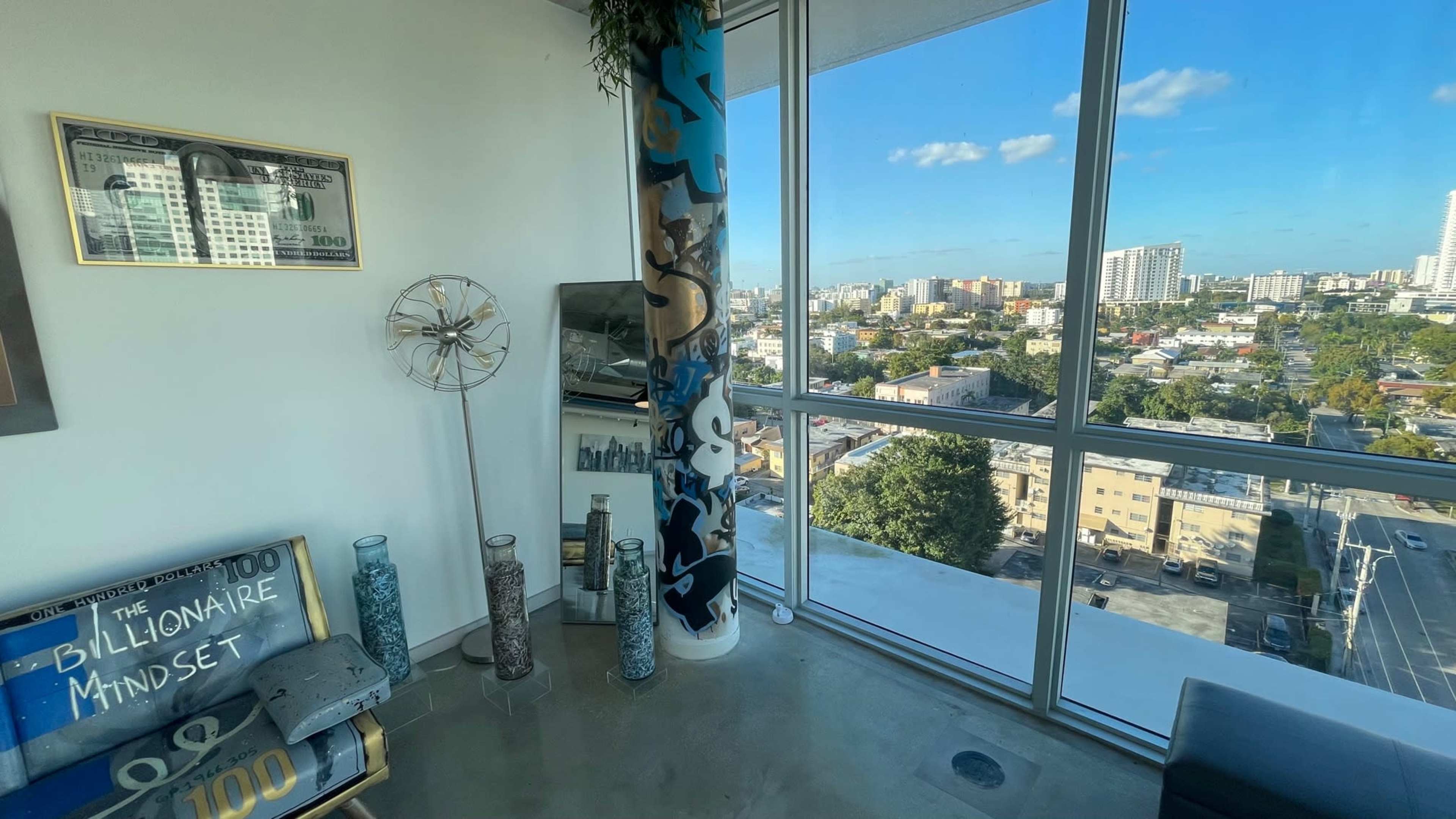 A modern room with large windows offering a view of a city skyline, complemented by decorative elements including a framed dollar bill and a "Billionaire Mindset" sign.