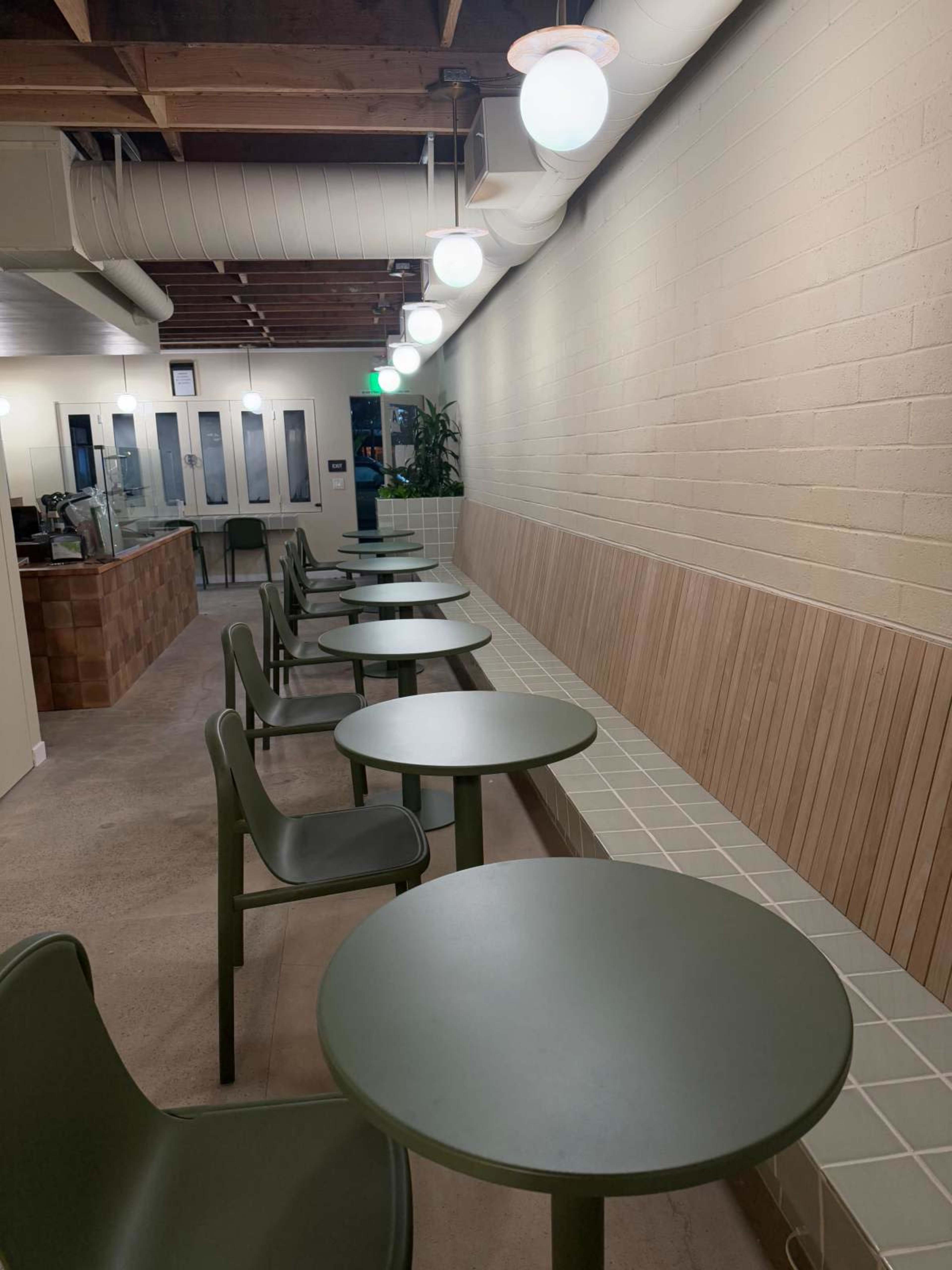 The image shows a line of green tables and chairs along a wall in a modern cafe with overhead lighting.