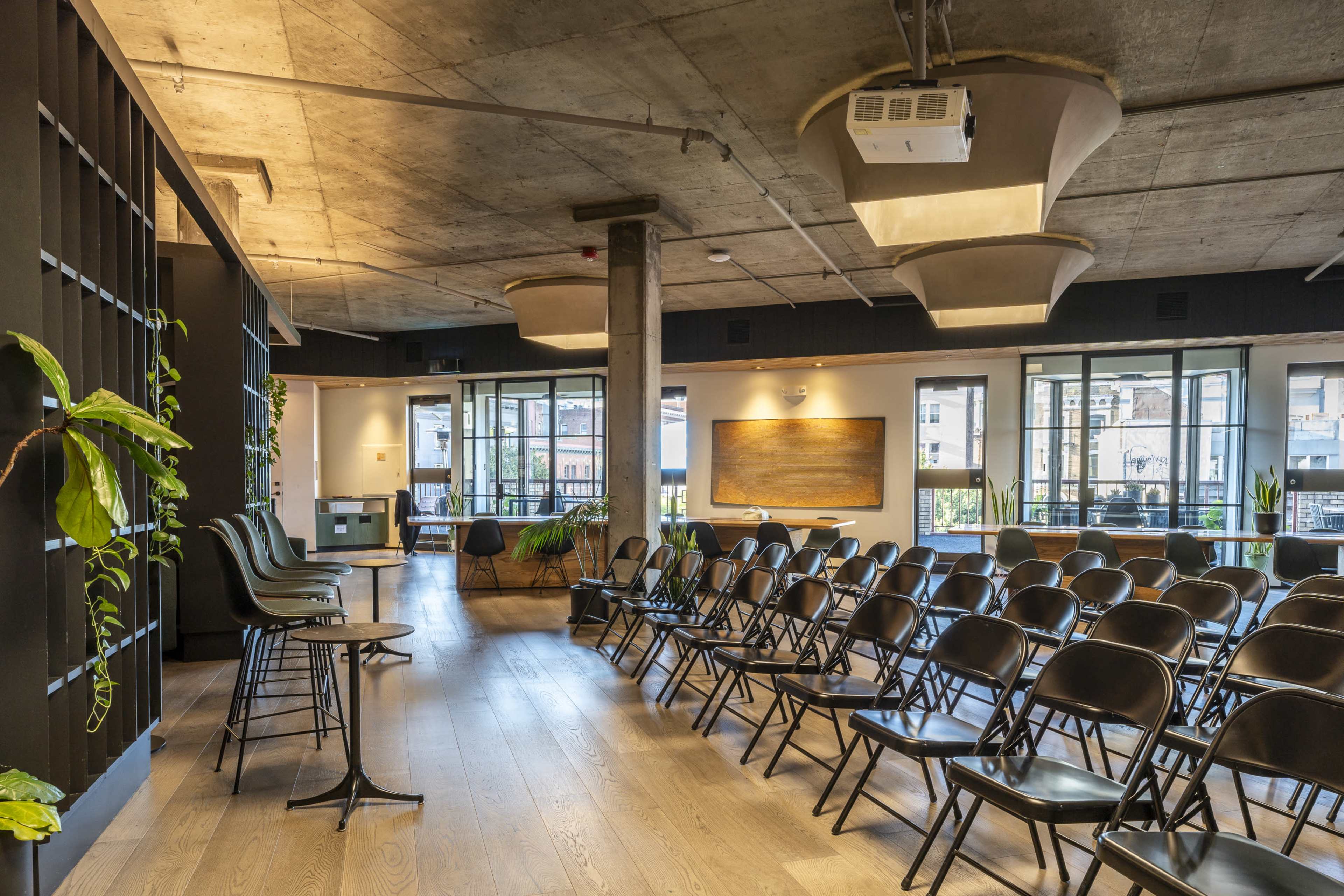 The image shows a modern conference room set up with rows of black folding chairs facing a projector screen, surrounded by large windows and greenery.