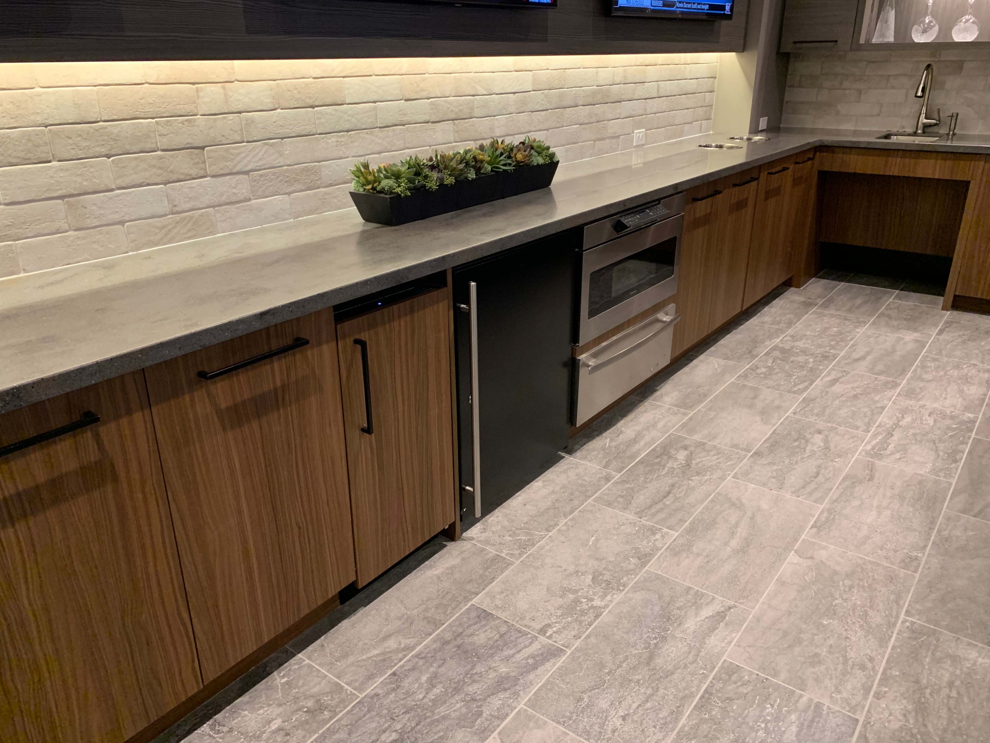 A modern kitchen with wooden cabinetry, a gray countertop, a built-in oven, and a row of small plants in a planter.