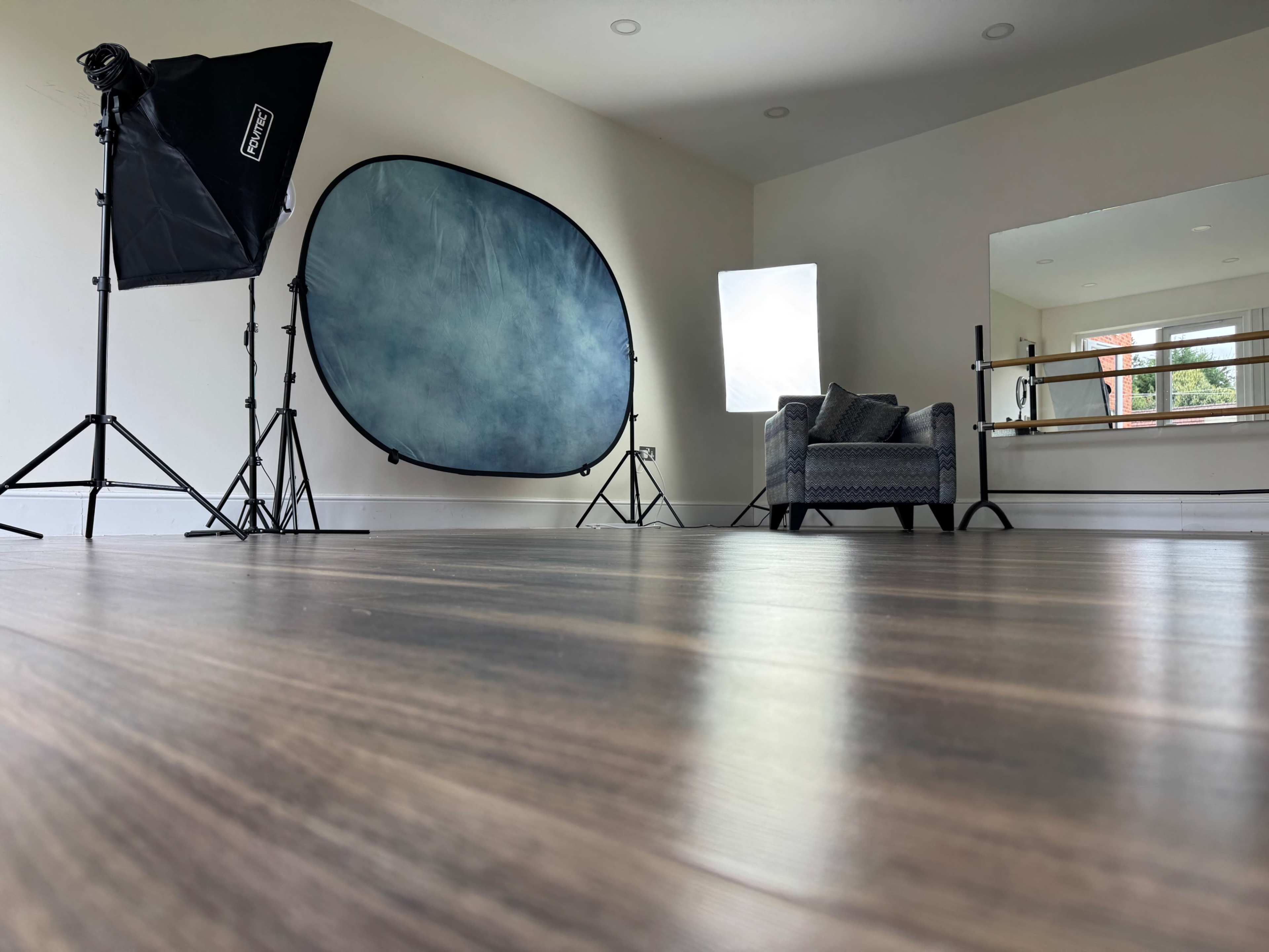 The image shows a photography studio with a backdrop, lighting equipment, and a single gray chair placed near a large mirror.
