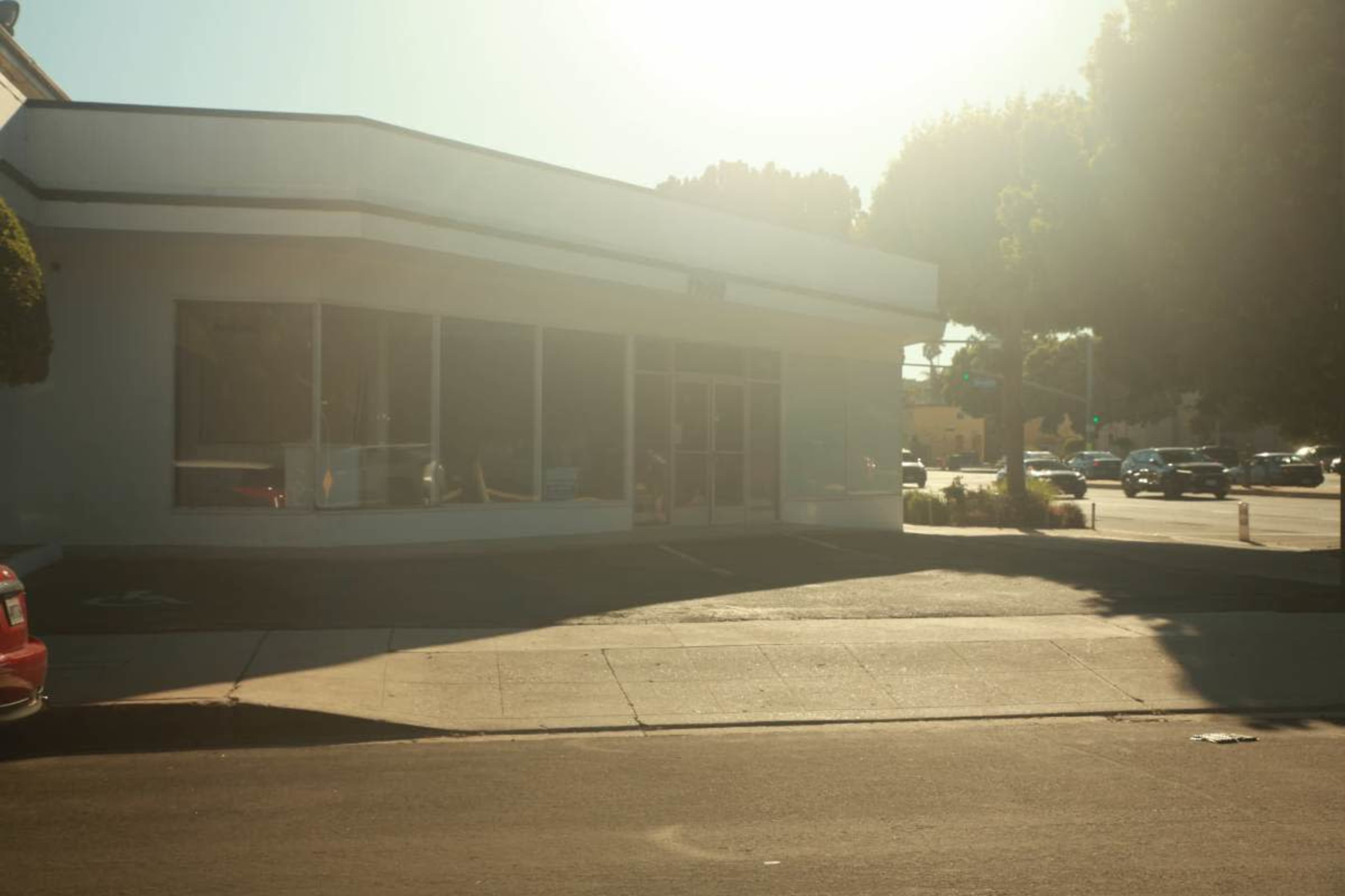 A modern single-story building with large windows is situated on a street corner, illuminated by bright sunlight.