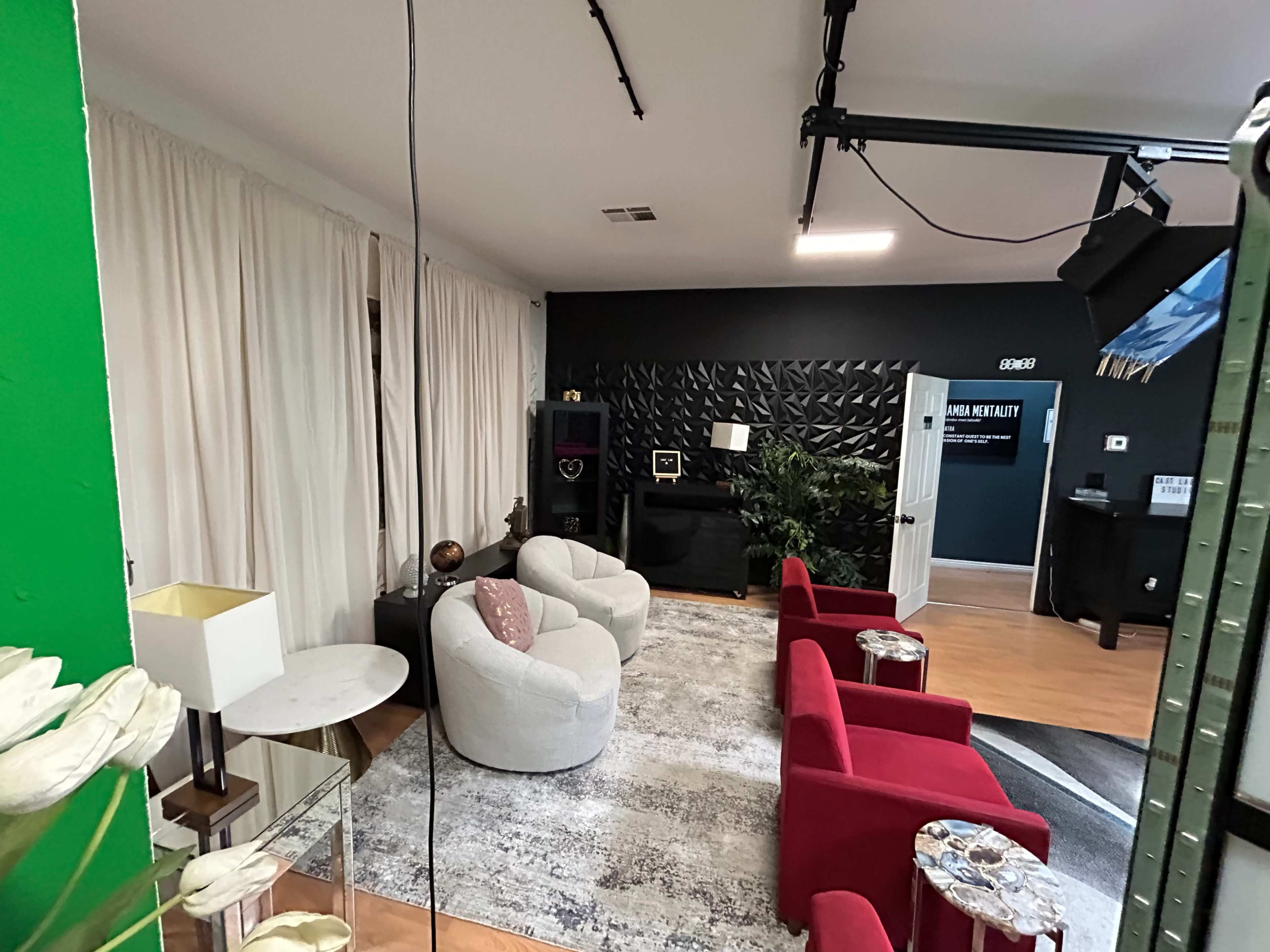 The image shows a modern interior space with a mix of seating options, including white and red chairs, a large area rug, and decorative plants against a dark wall.