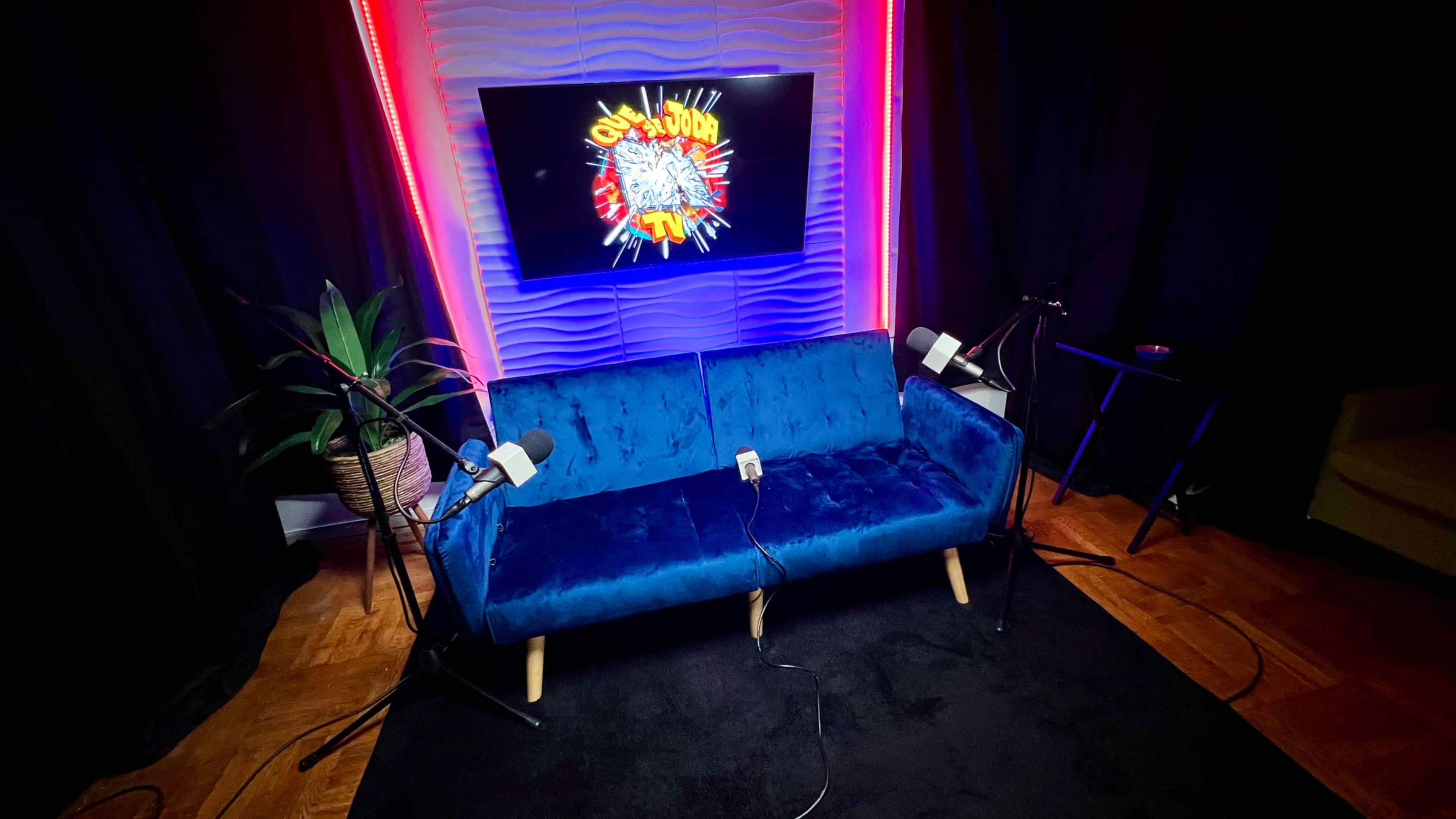 The image shows a blue velvet couch placed in a studio setting with two microphones on stands and a brightly lit screen displaying a colorful logo in the background.