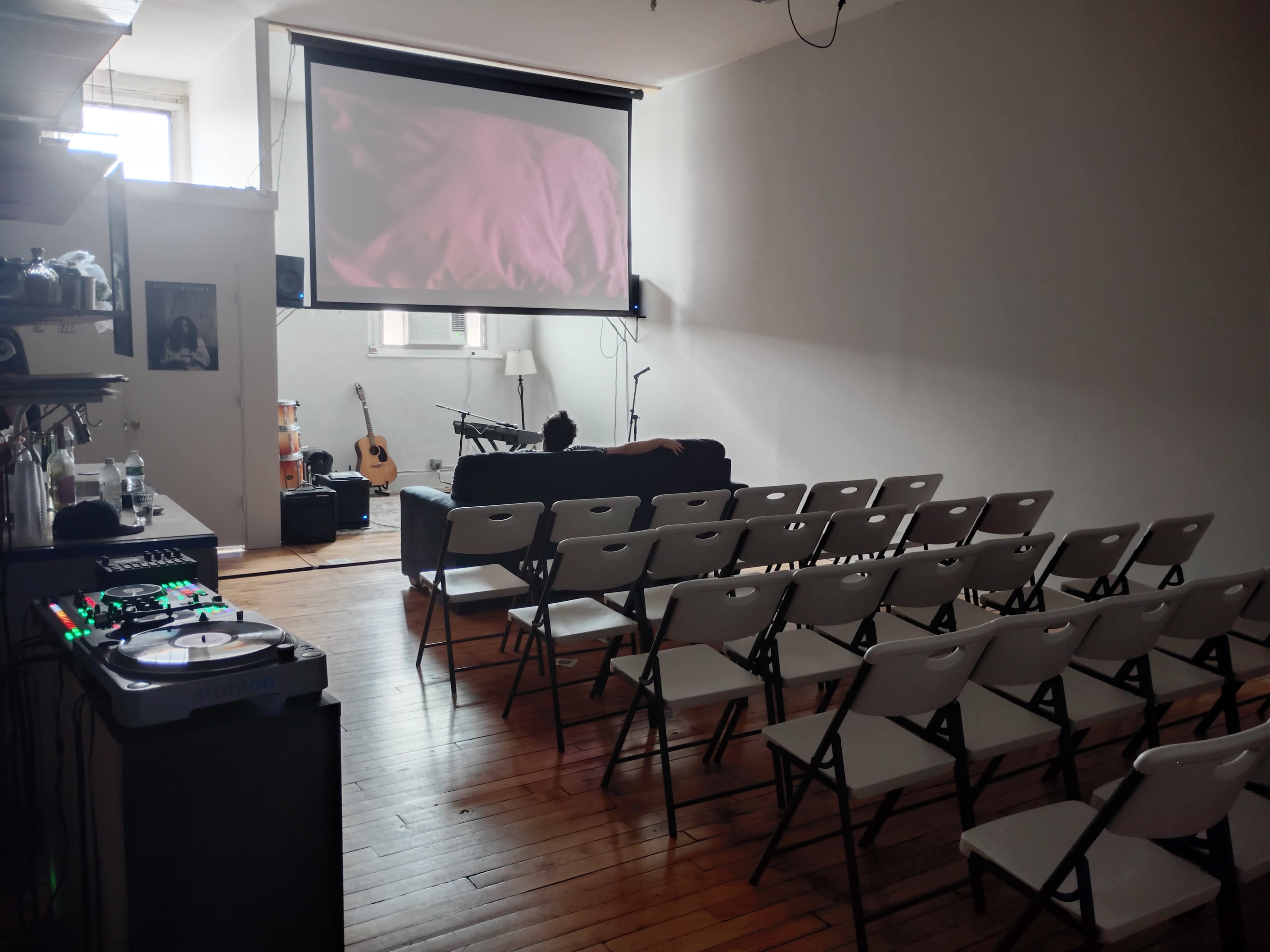 The image shows a compact indoor venue with folding chairs arranged in rows facing a large projection screen, and a couch in the background alongside musical instruments.