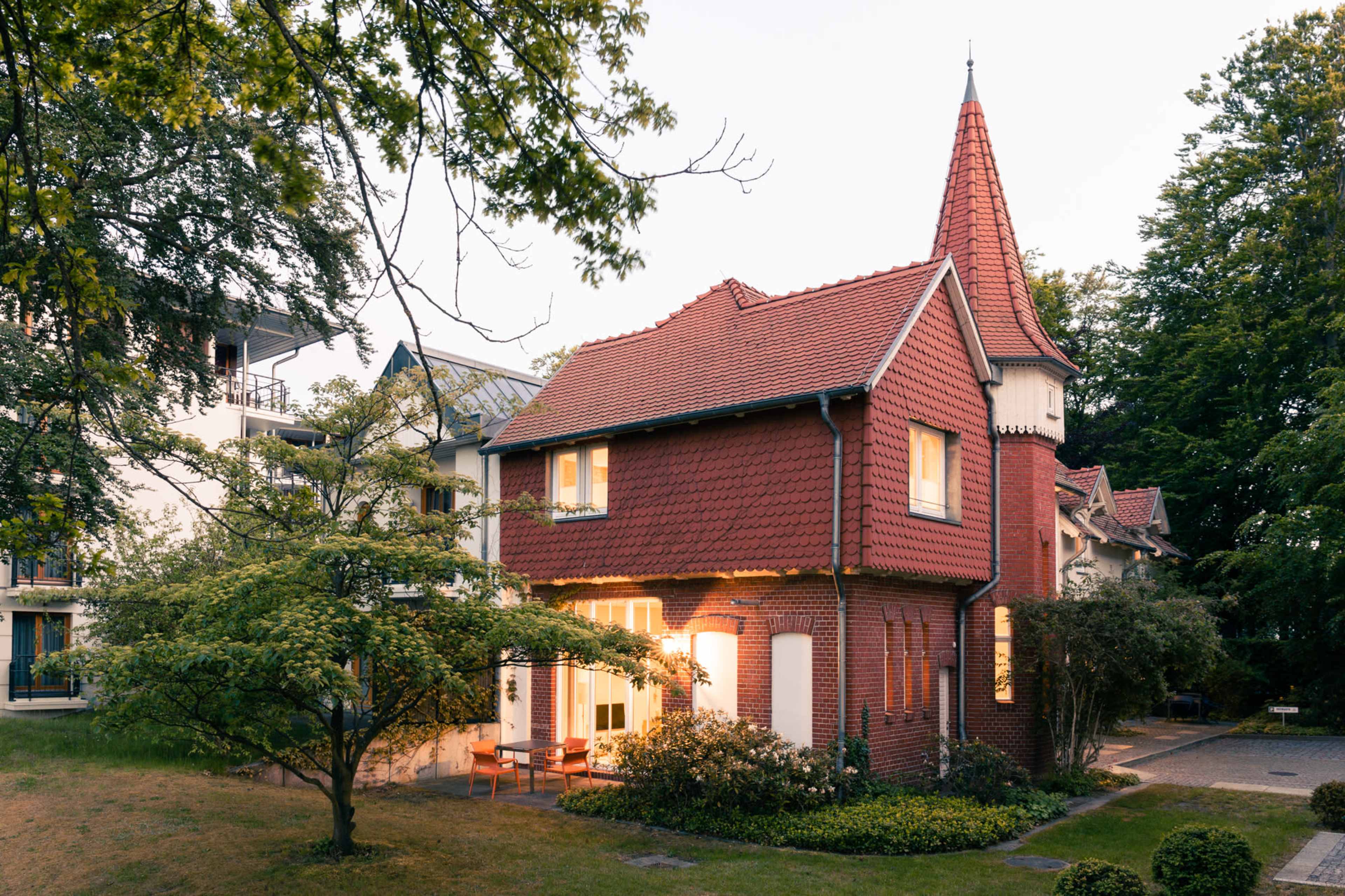 A red-roofed house with a pointed tower is surrounded by trees and greenery.