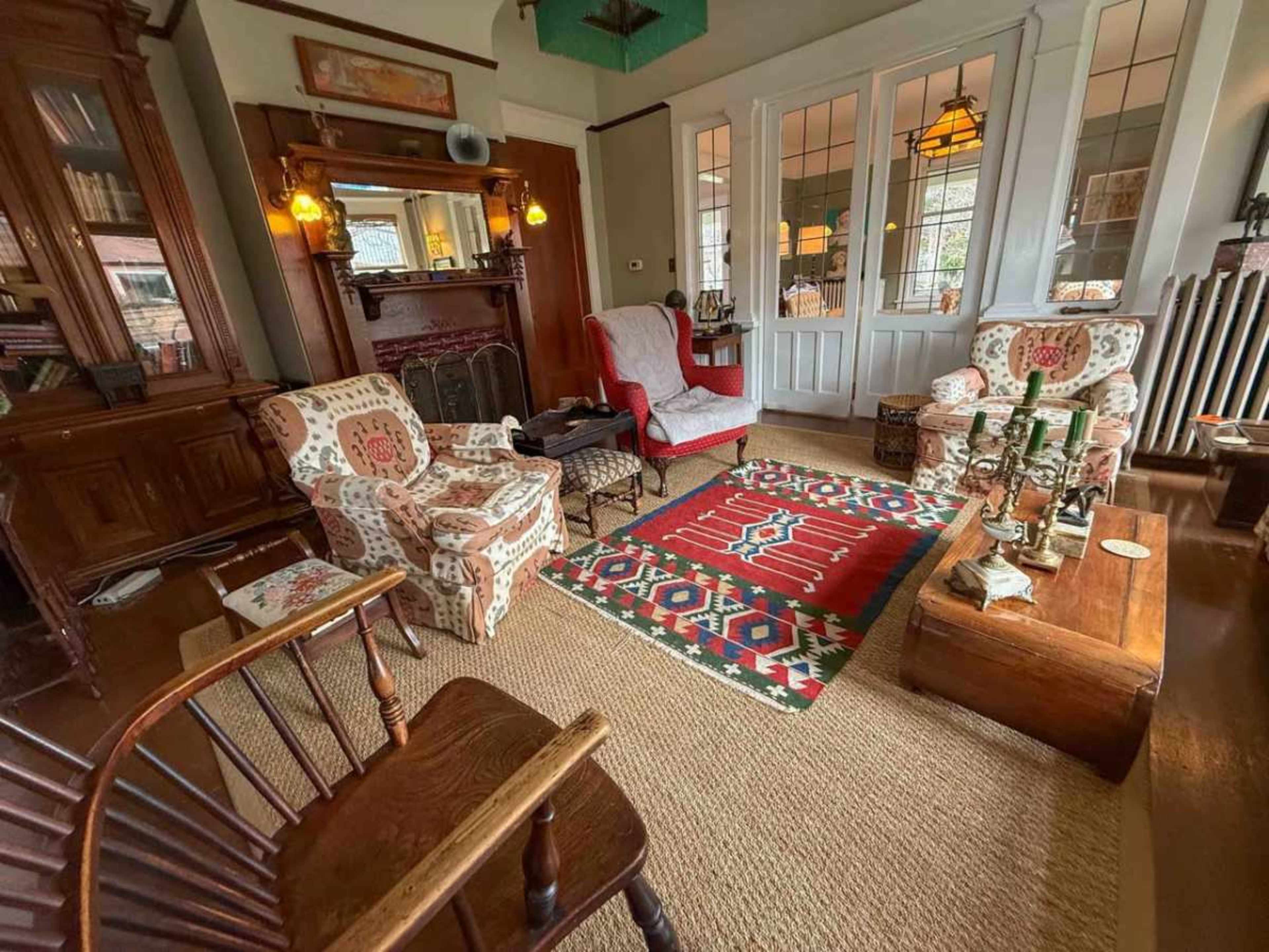 The room features two patterned armchairs, a wooden coffee table, and a large area rug on a hardwood floor, with bookshelves and a decorative fireplace in the background.