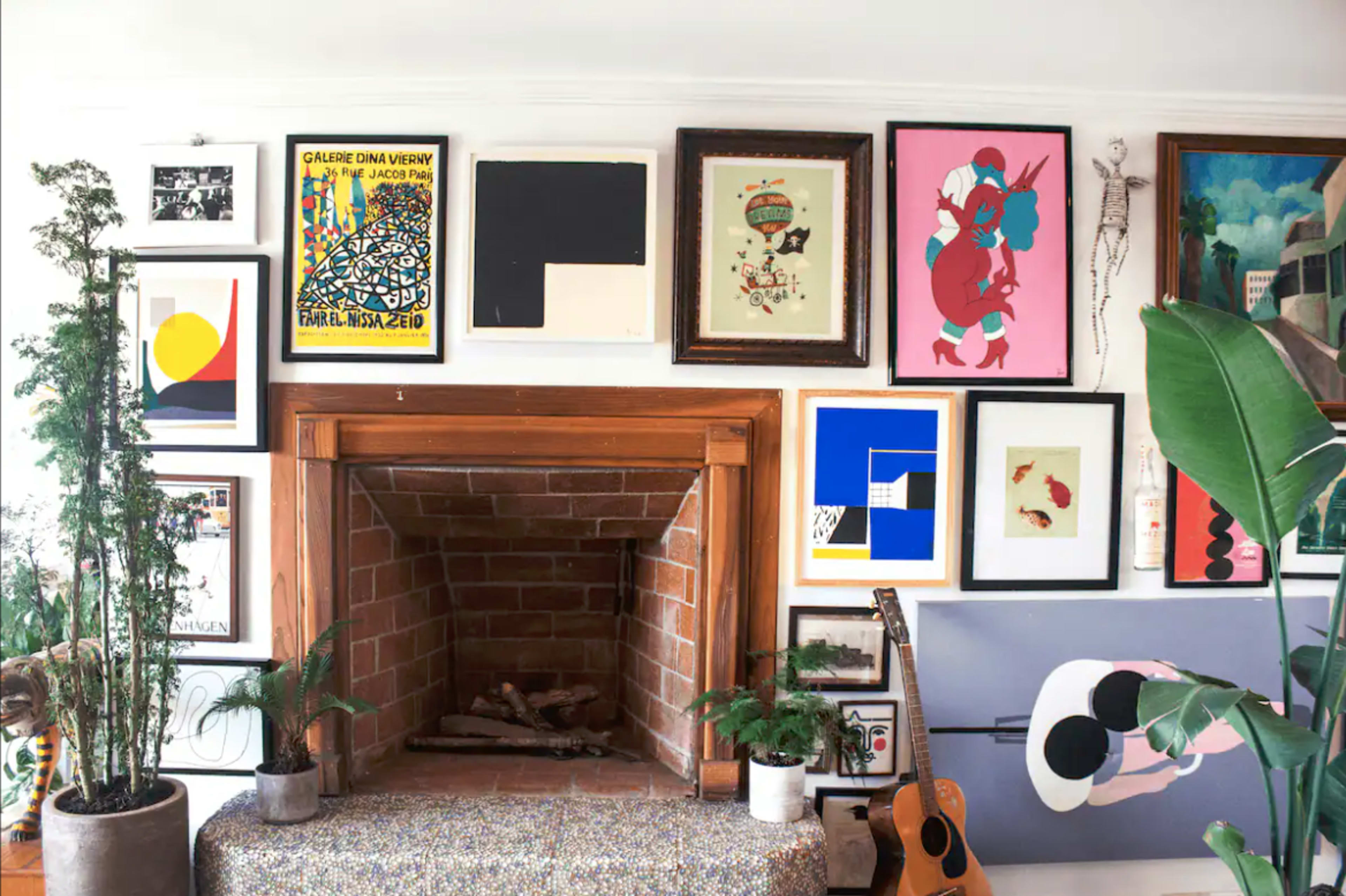 A living room wall with a brick fireplace surrounded by a diverse collection of framed artwork and two potted plants.