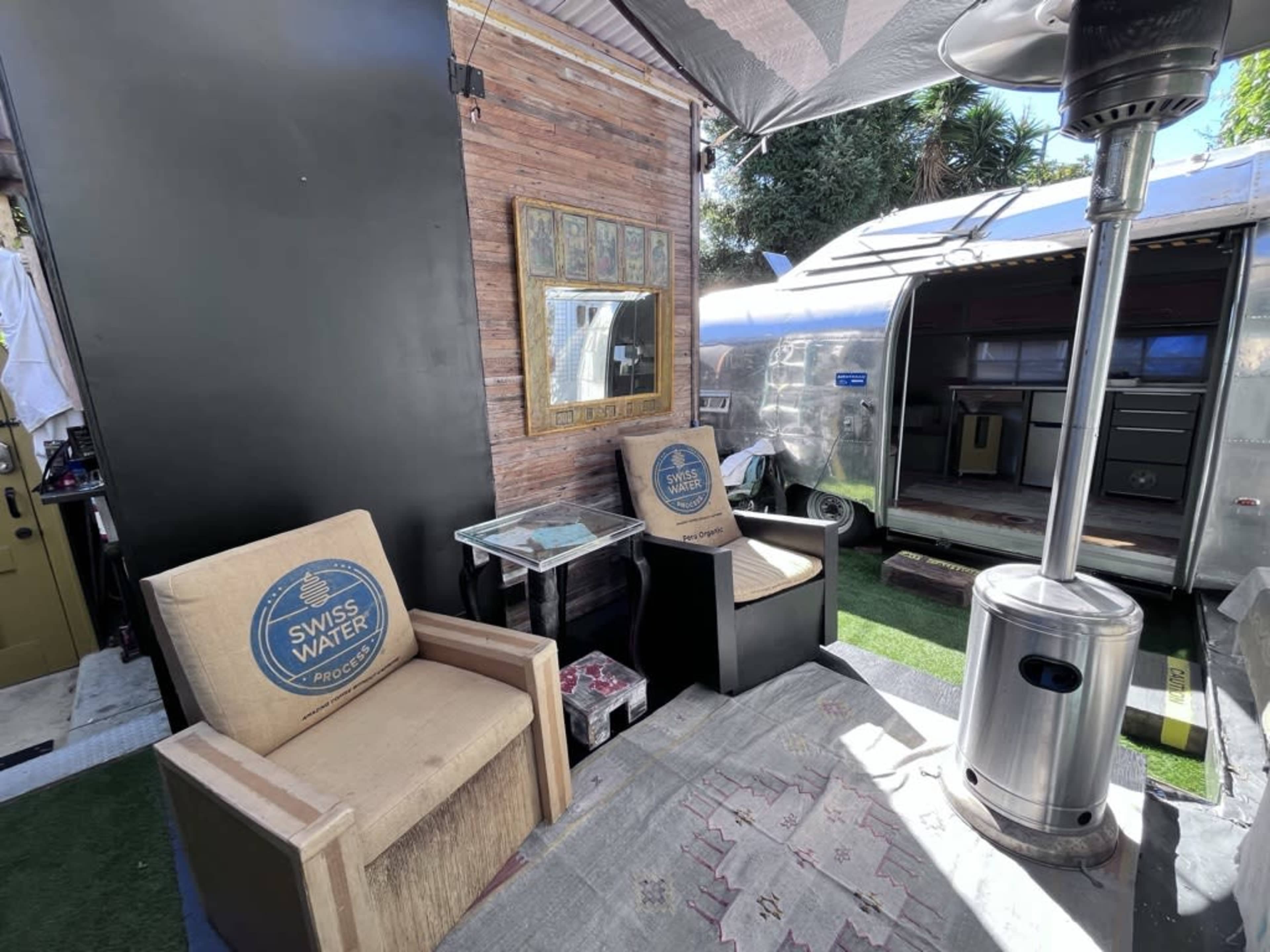 A seating area with two chairs and a small table next to an Airstream trailer under a shaded canopy.