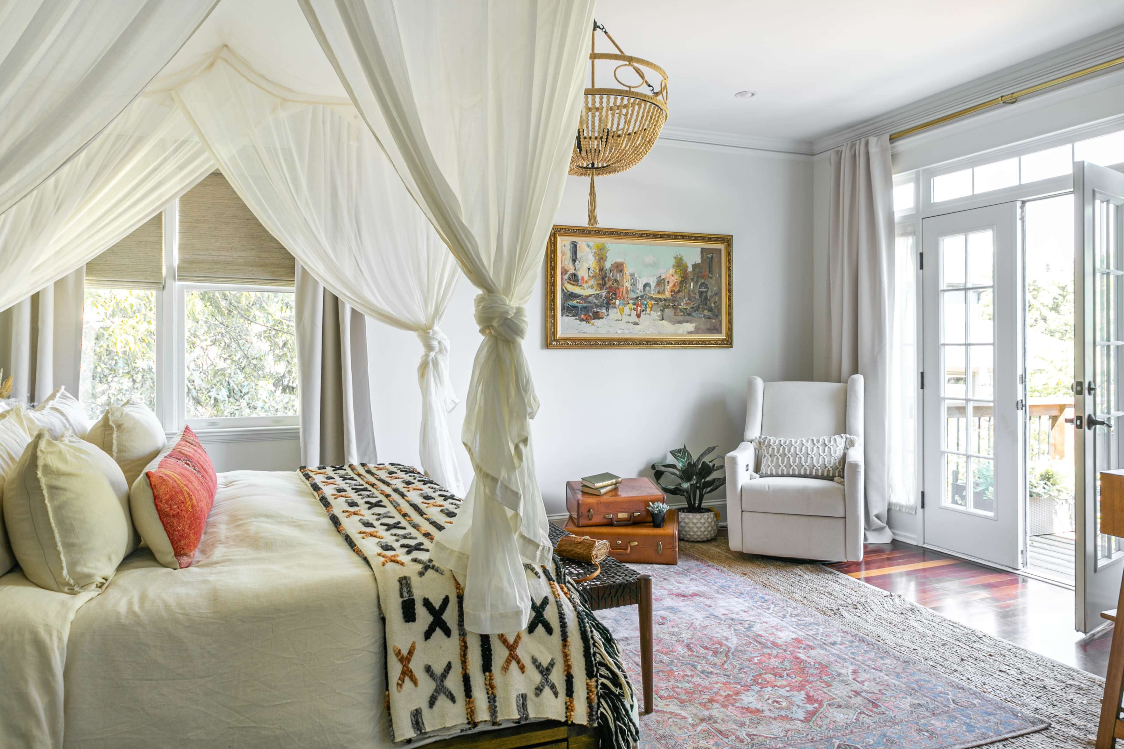 A light-filled bedroom features a canopied bed, a large artwork on the wall, a comfortable seating area, and a door leading to an outdoor space.