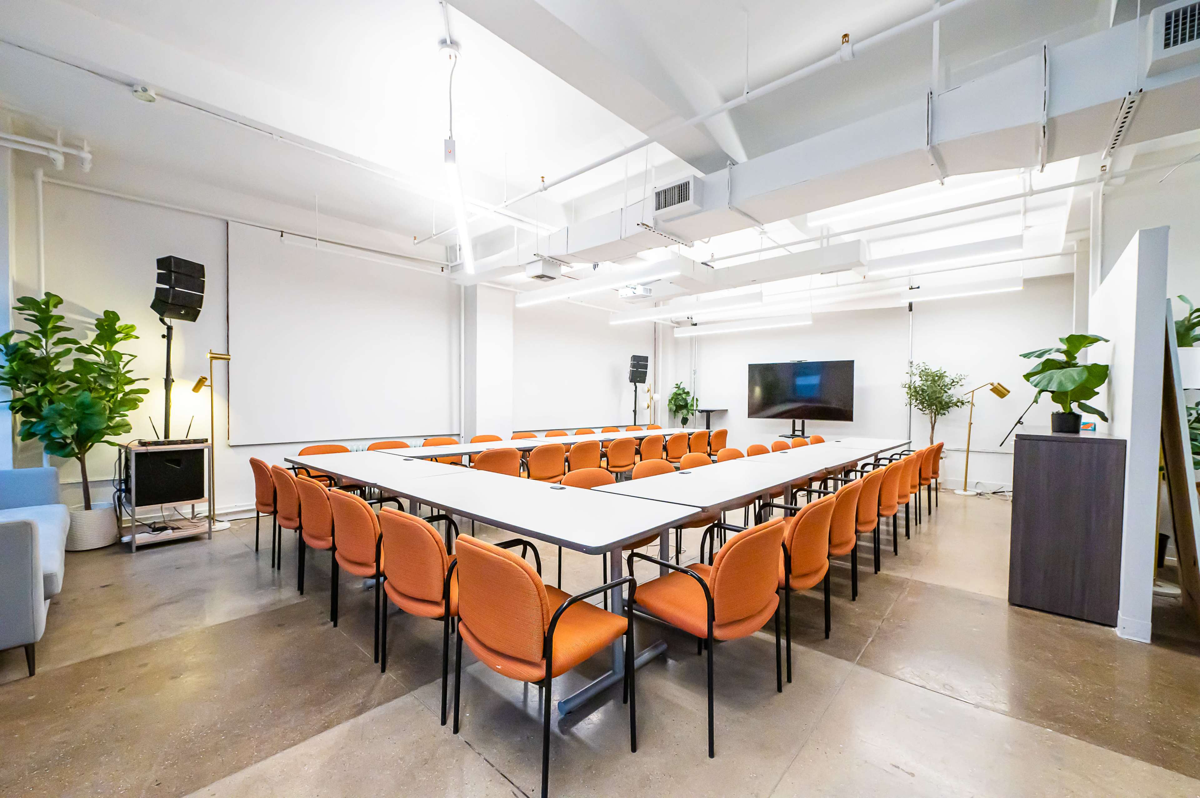 A conference room is arranged with orange chairs surrounding a long rectangular table, accompanied by plants and a large screen at one end.