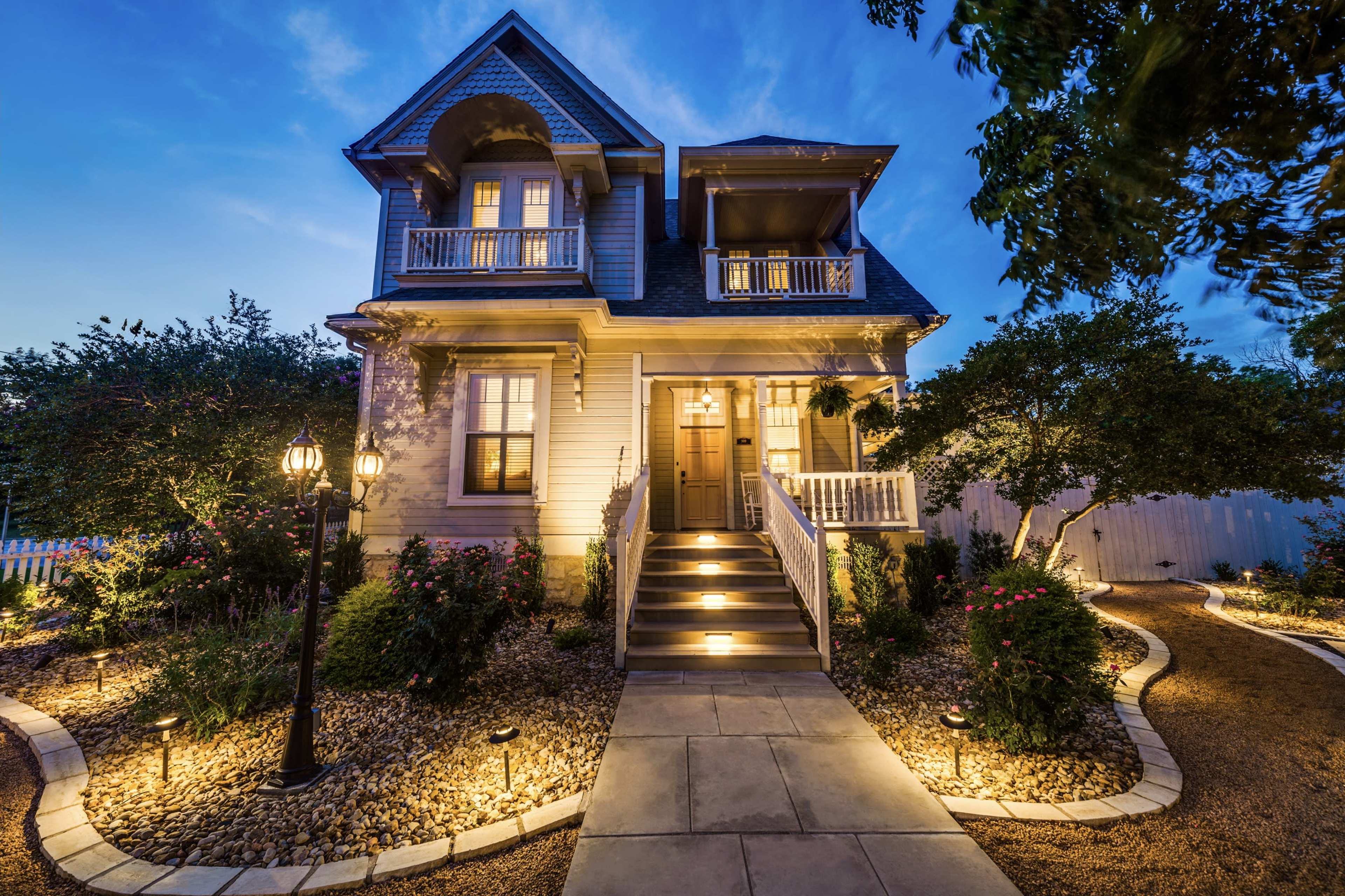 A well-lit two-story house with a porch and landscaped garden is seen at twilight.