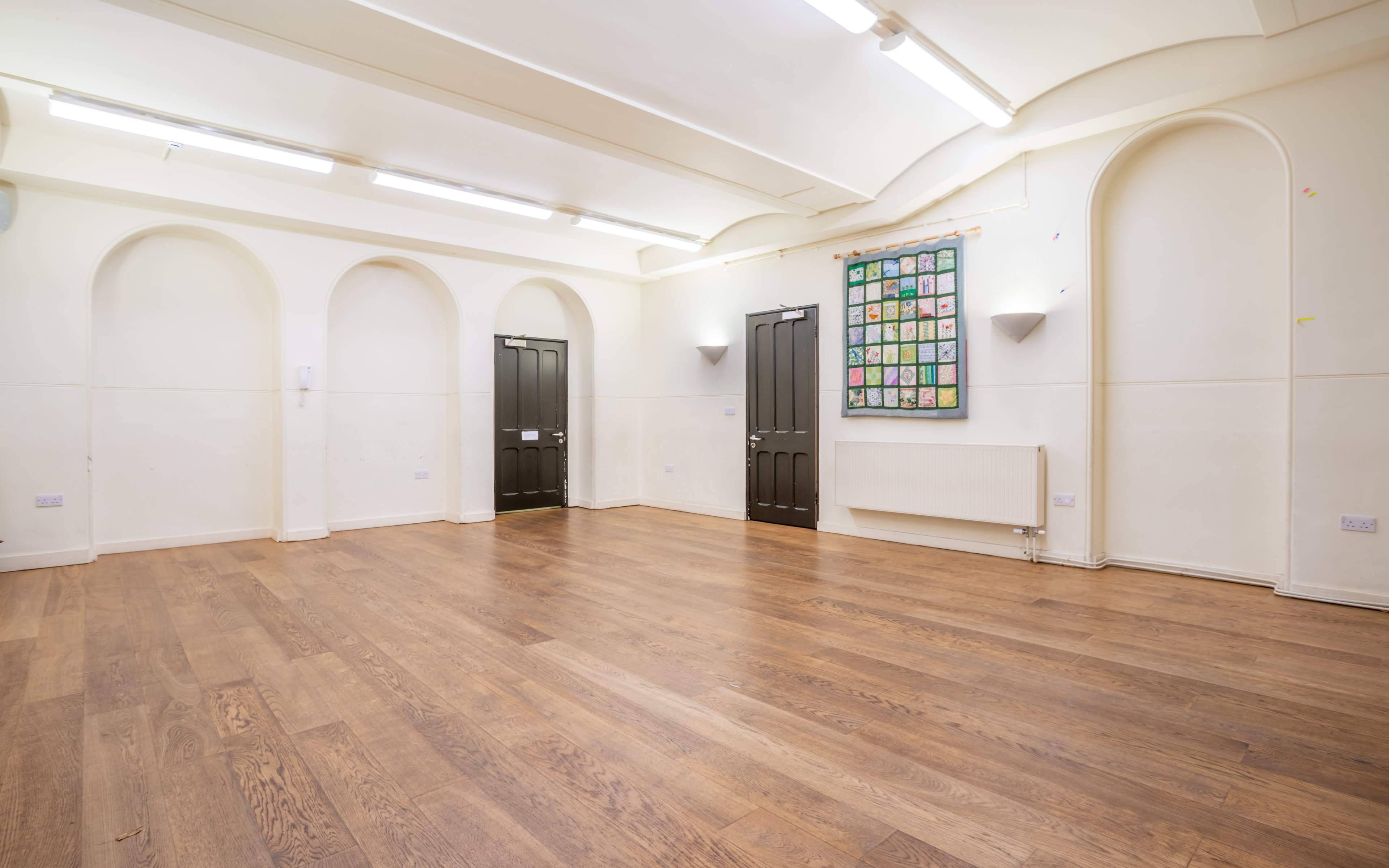 An empty room with wooden flooring, white walls, and two black doors, along with a wall-mounted radiator and a framed collection of artwork.
