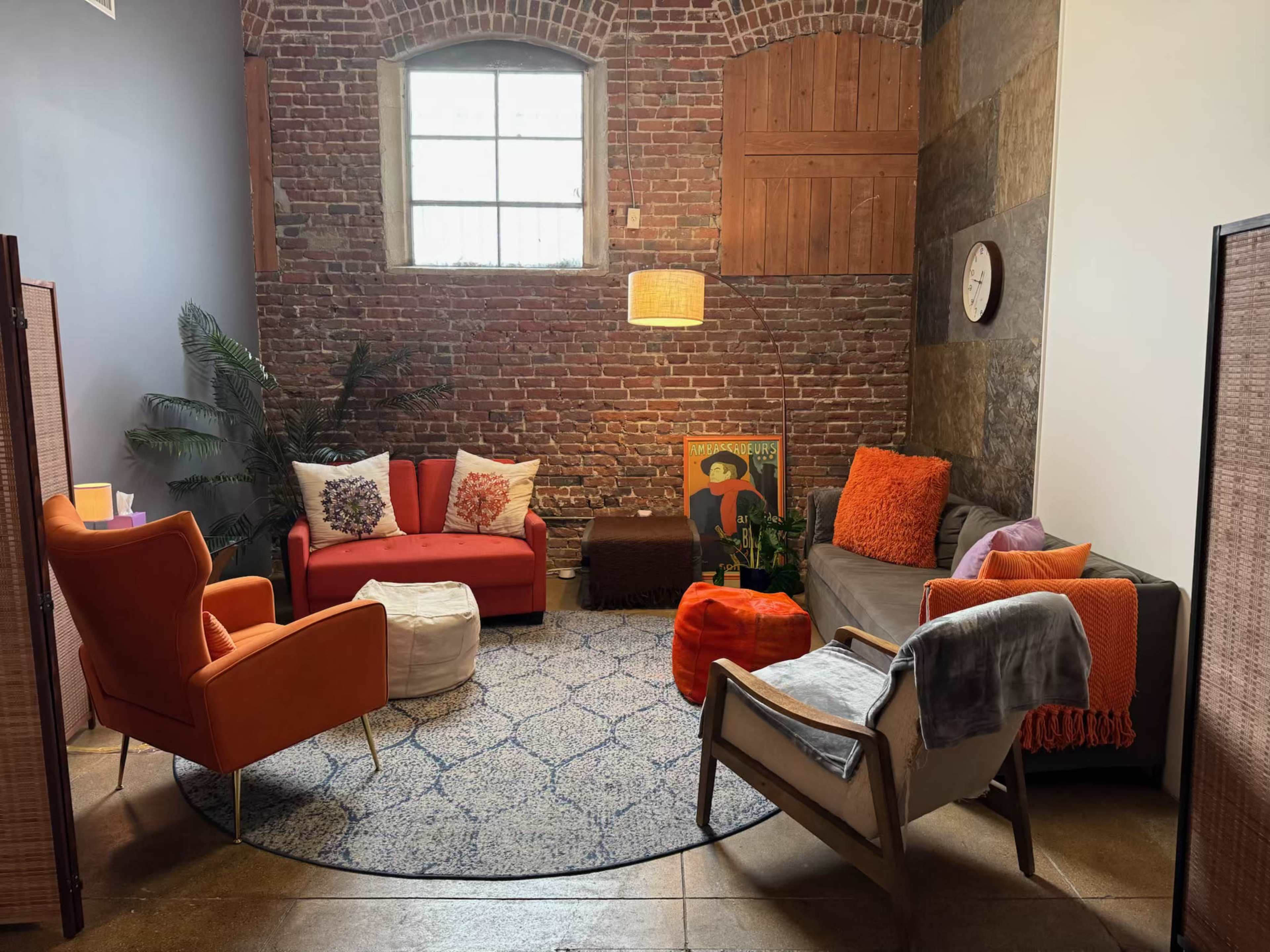 The image shows a cozy lounge area with brick walls, various colorful seating options, a round rug, and a lamp hanging from the ceiling.