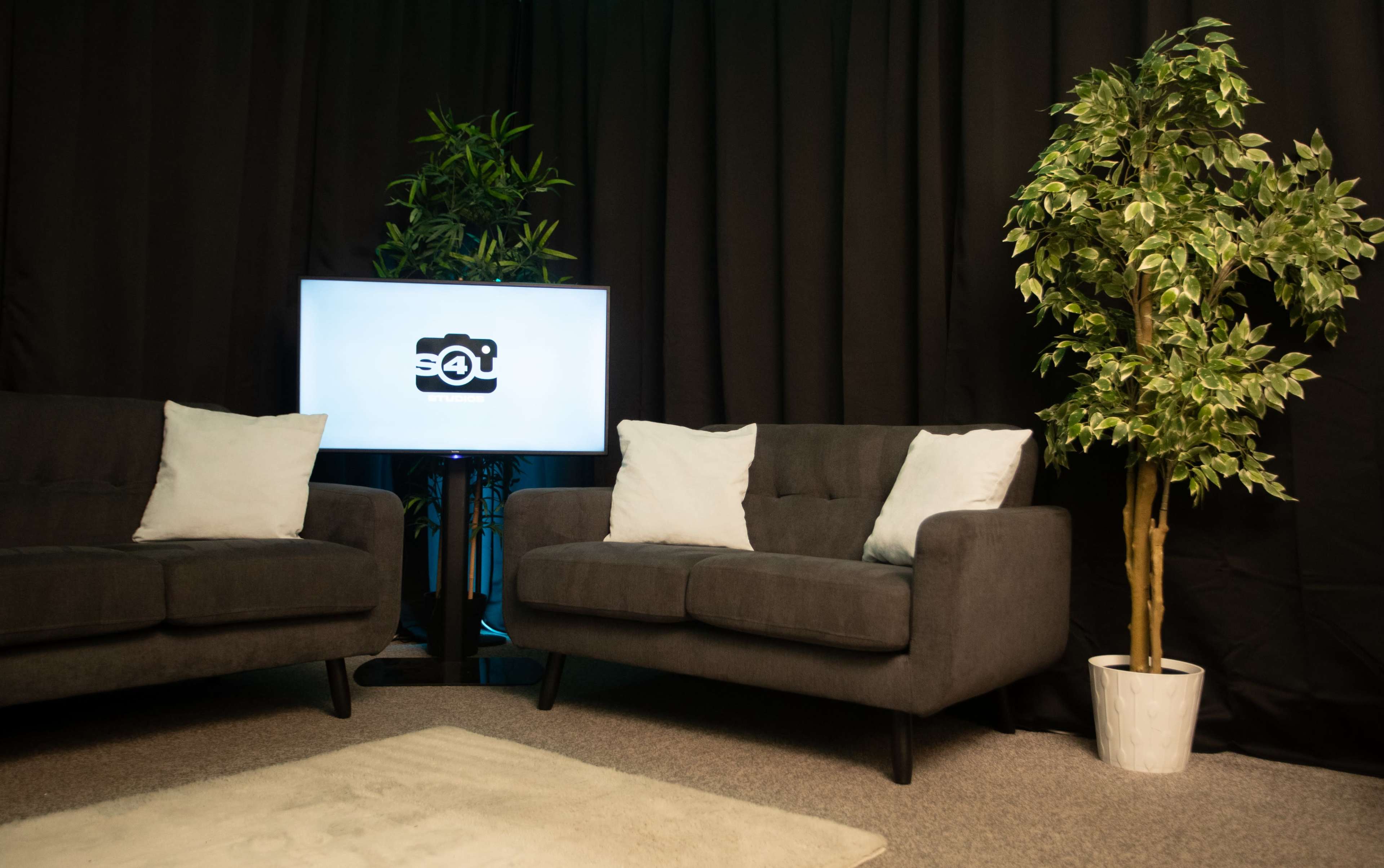 A cozy seating area with two gray sofas and a television screen displaying a logo, set against a black backdrop and accompanied by potted greenery.