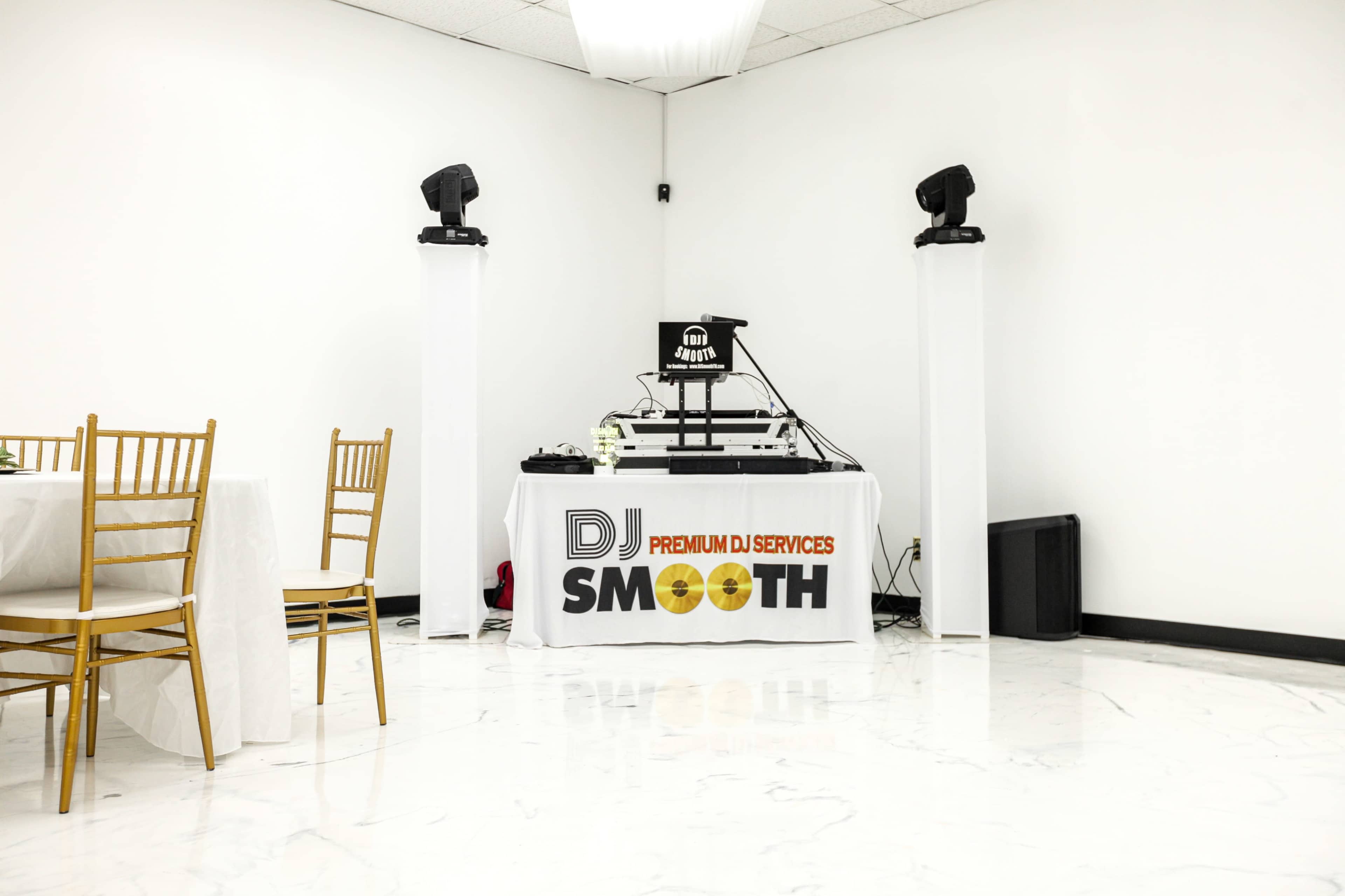 A DJ setup with speakers and a mixer is positioned in a corner of a white-walled room, accompanied by gold-framed tables.
