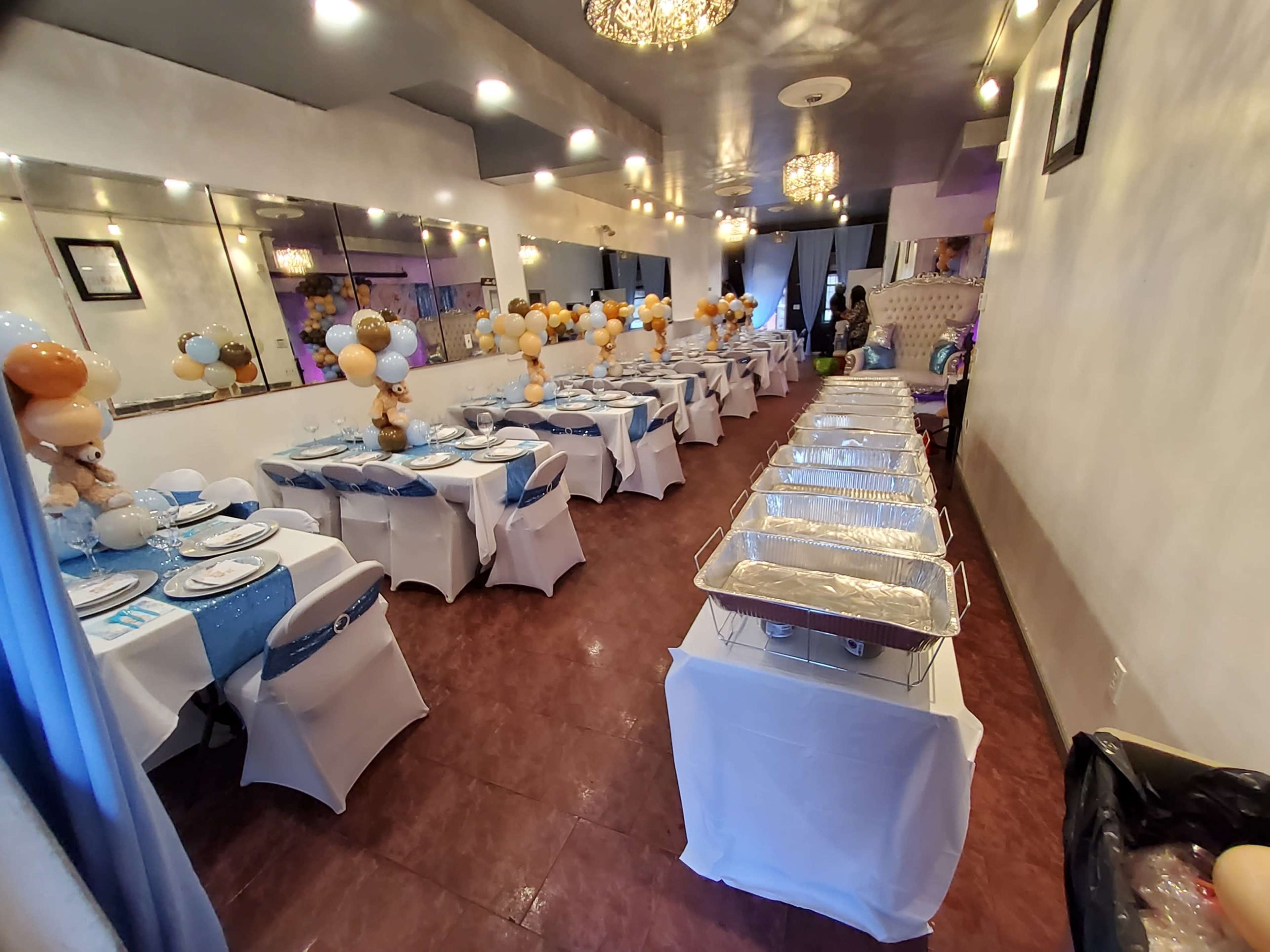 A banquet hall set up with long tables covered in white tablecloths and decorated with balloons, with a buffet area on one side.