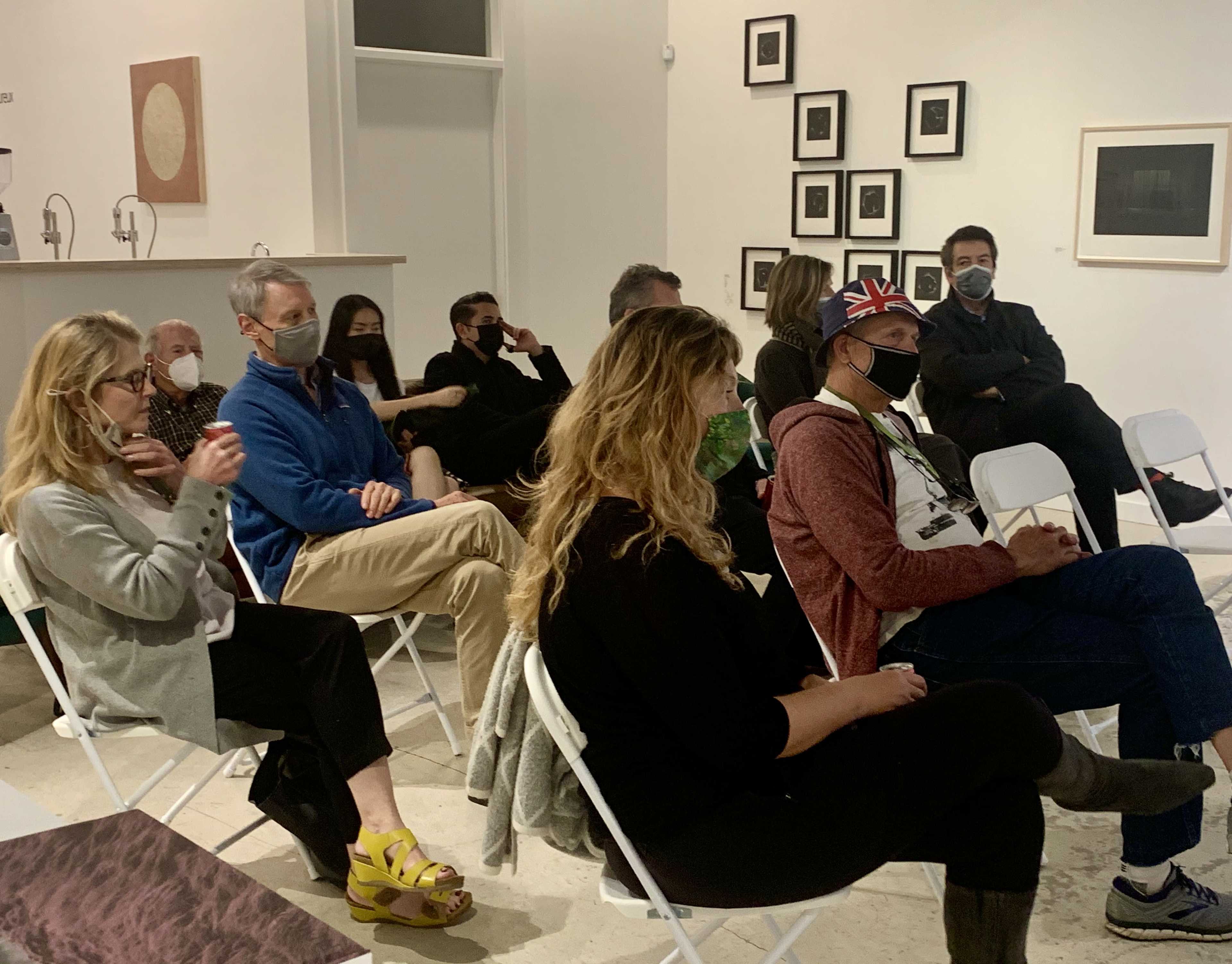A group of eight individuals wearing masks sits in folding chairs in a gallery setting, while one person stands in the foreground.