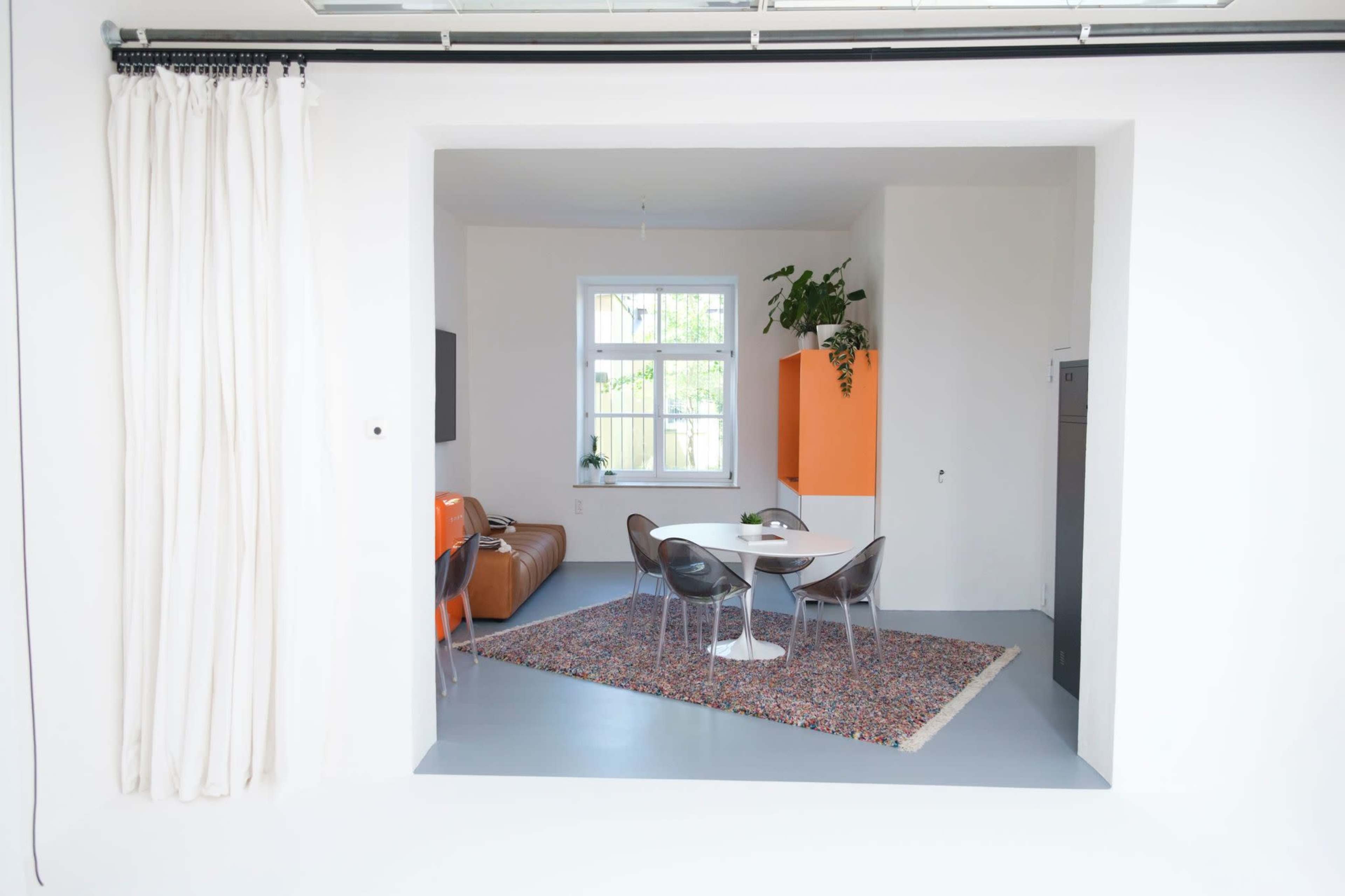 The image shows a minimalist interior space featuring a dining area with a round table and chairs, surrounded by light-colored walls and orange accents.