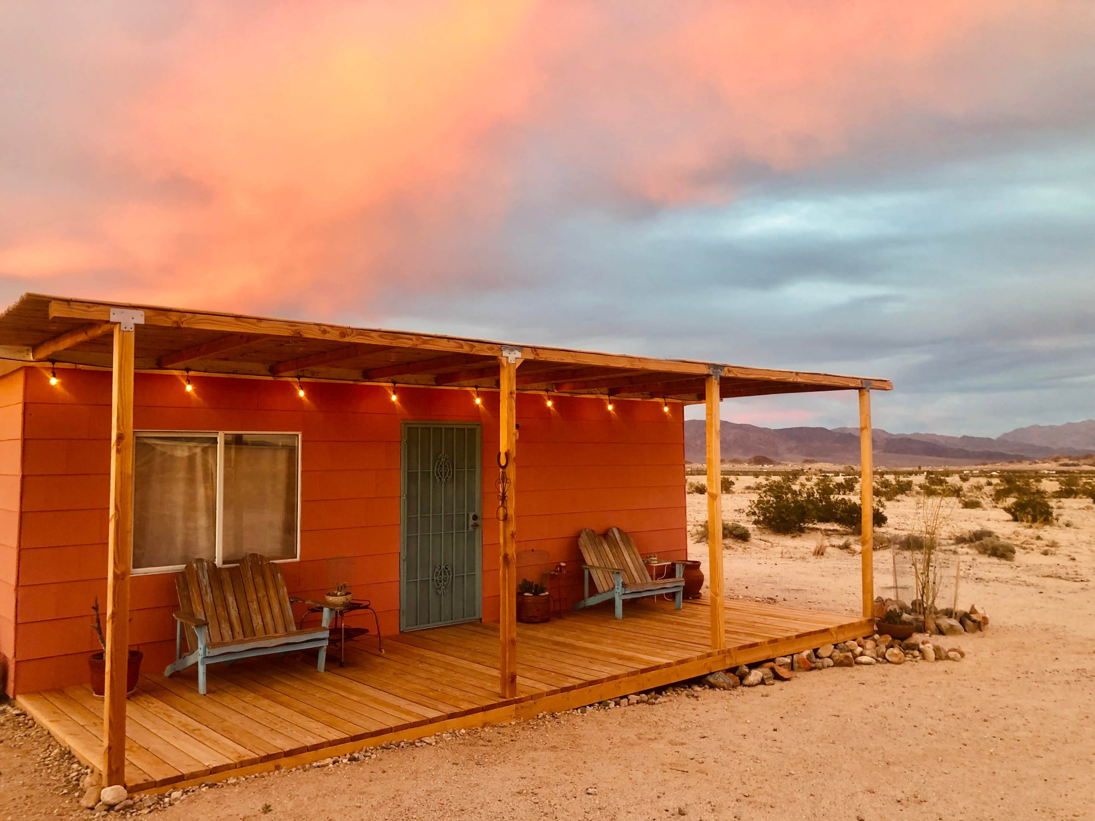A small orange cabin with a porch and blue chairs is surrounded by desert terrain under a colorful sky.