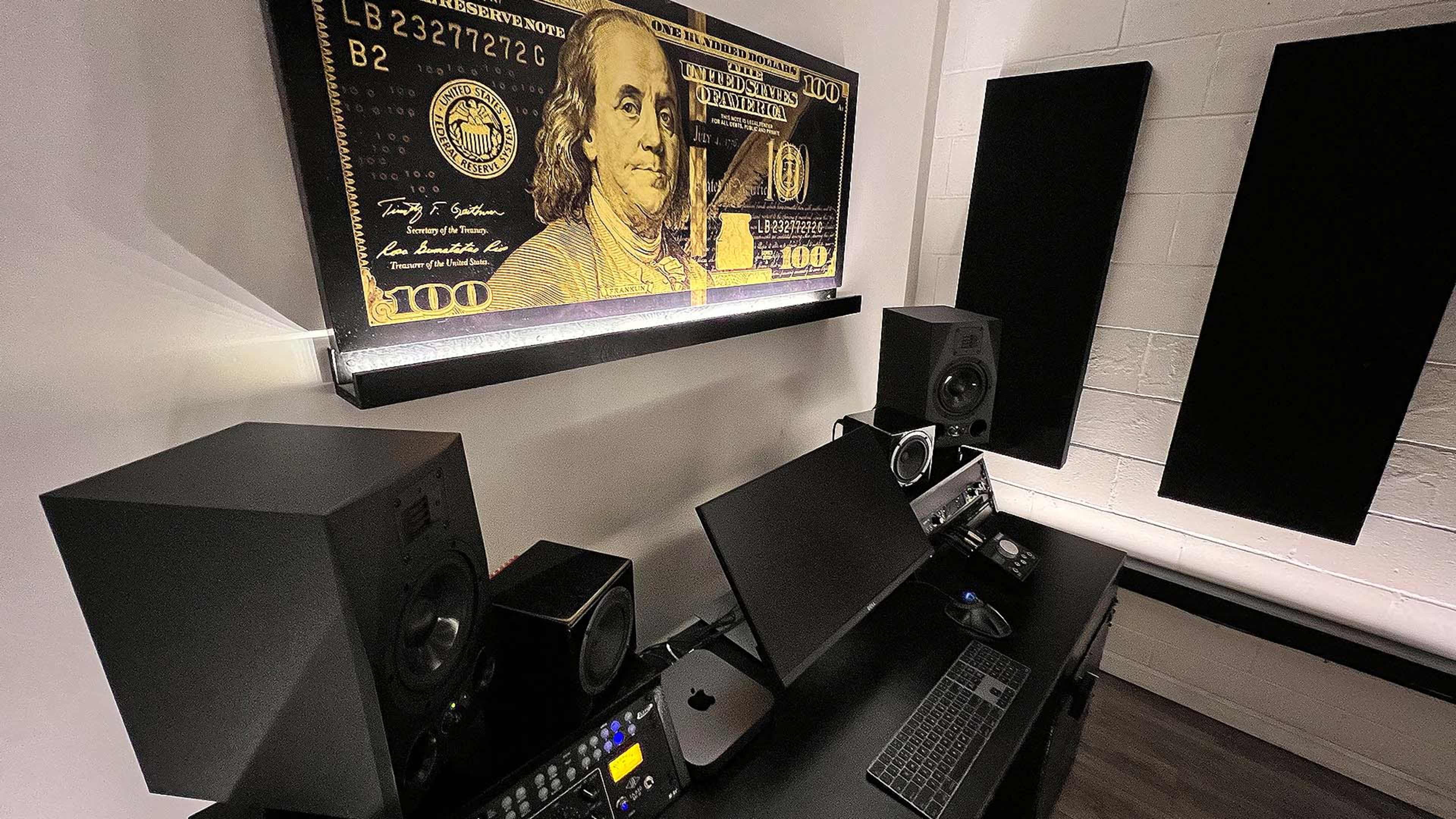 A music production studio featuring a large decorative $100 bill artwork above a sleek black workstation with speakers and a computer setup.