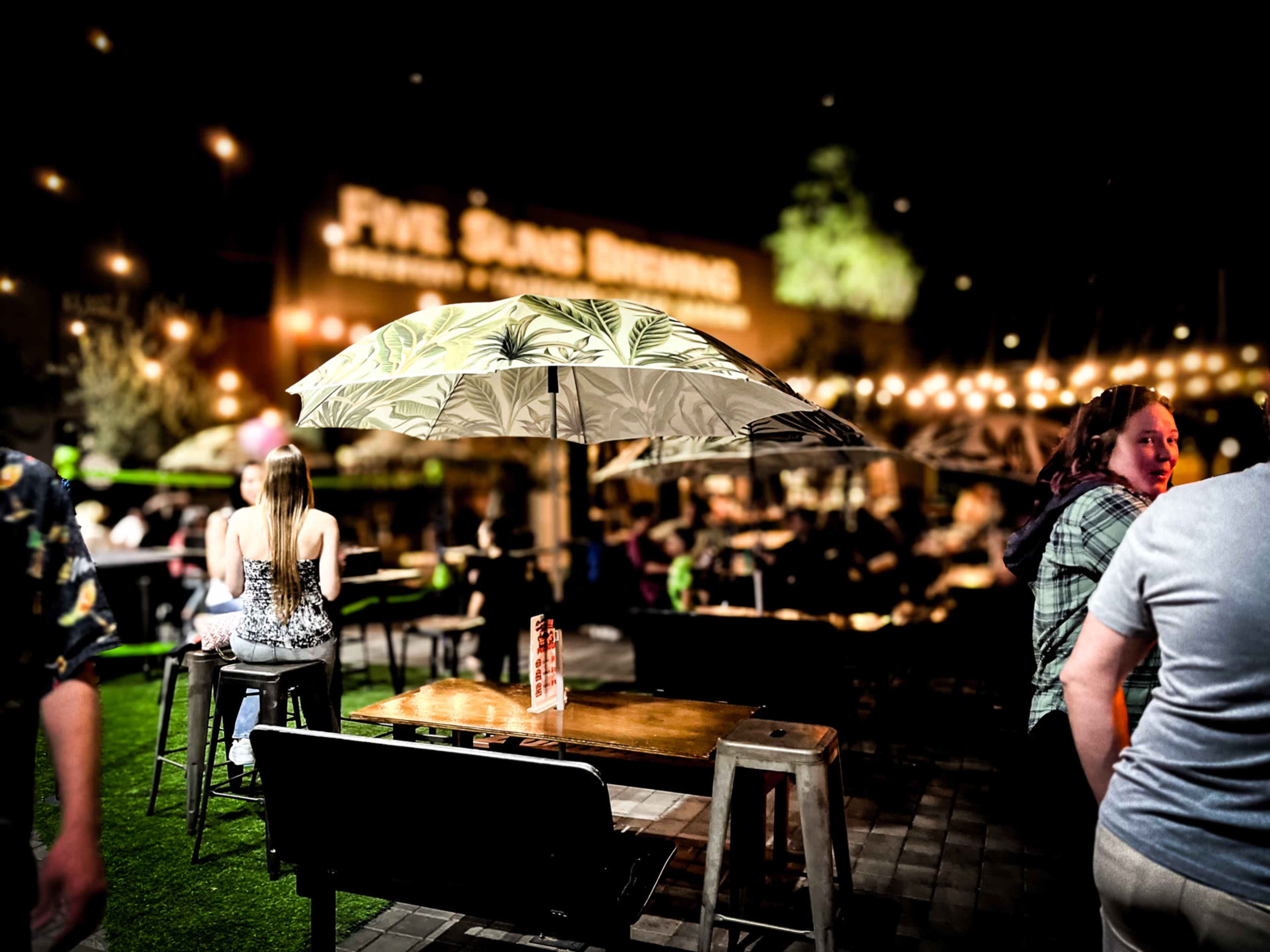 A lively outdoor dining area with tables and umbrellas, illuminated by string lights at night.