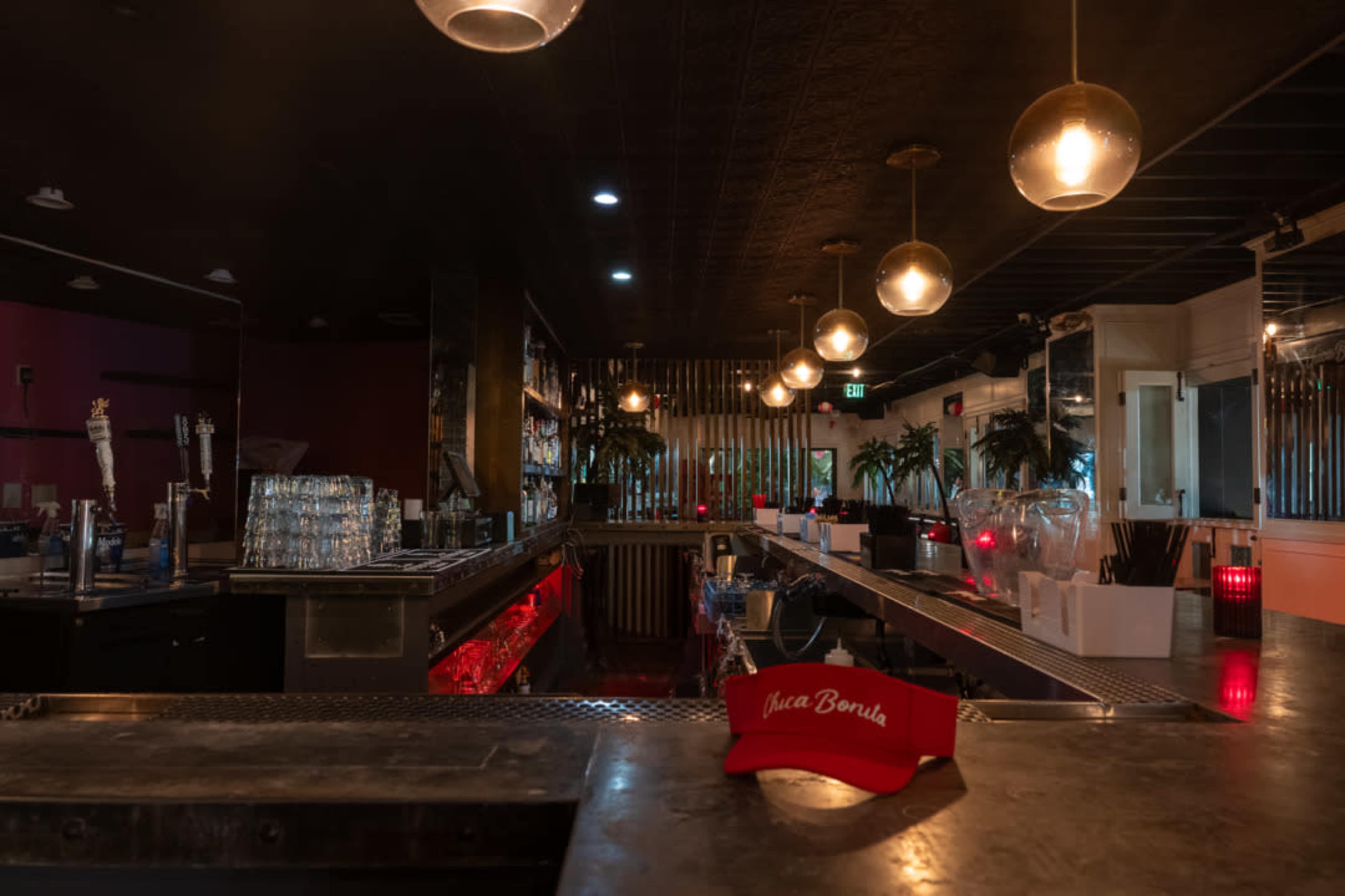 The image shows a dimly lit bar interior with a metal counter, illuminated by hanging light fixtures, and a red hat featuring the text "Casa Bonita" placed prominently in the foreground.