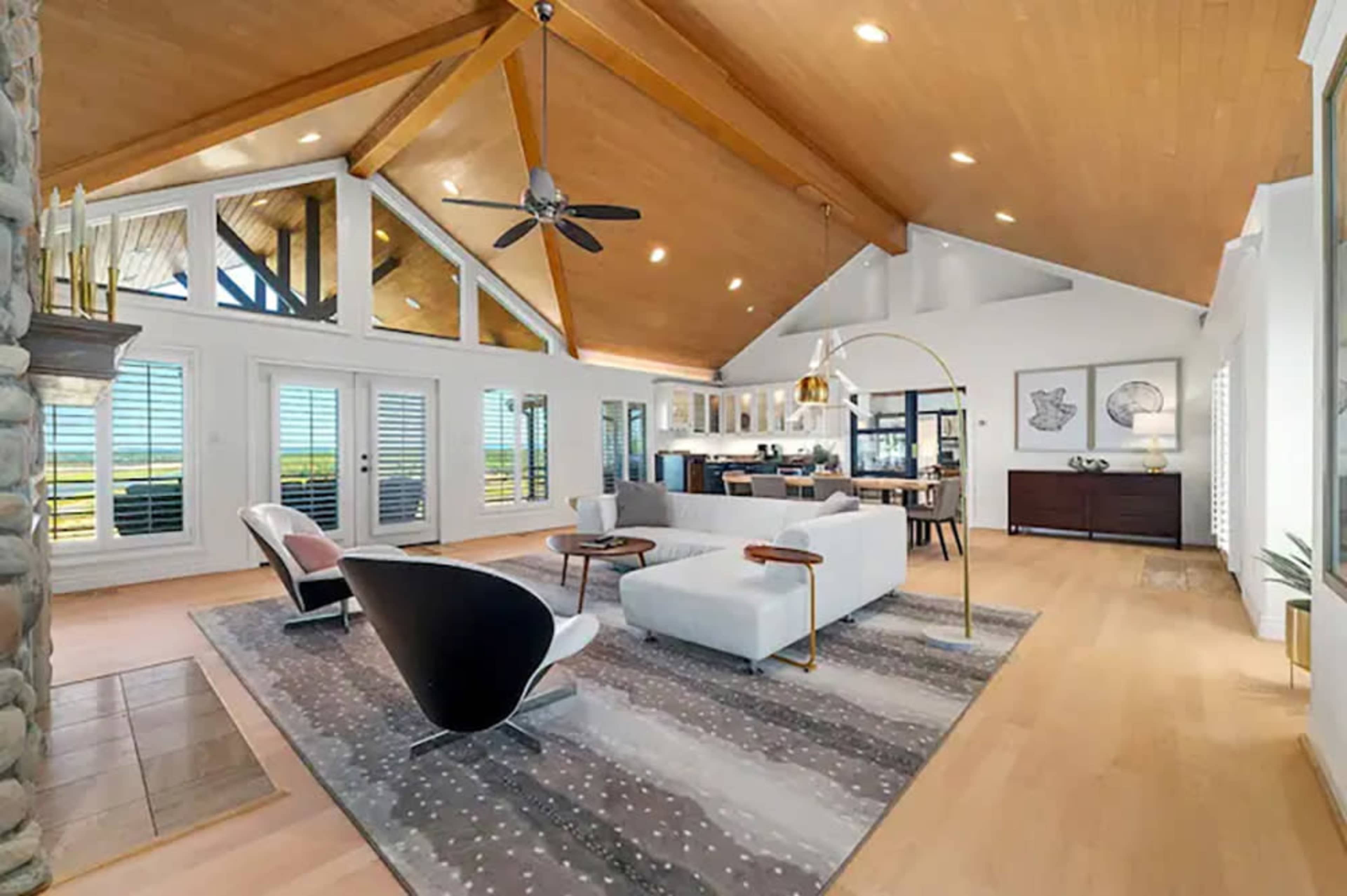 The image shows a spacious, modern living room with high ceilings, large windows, and a mix of contemporary furniture.