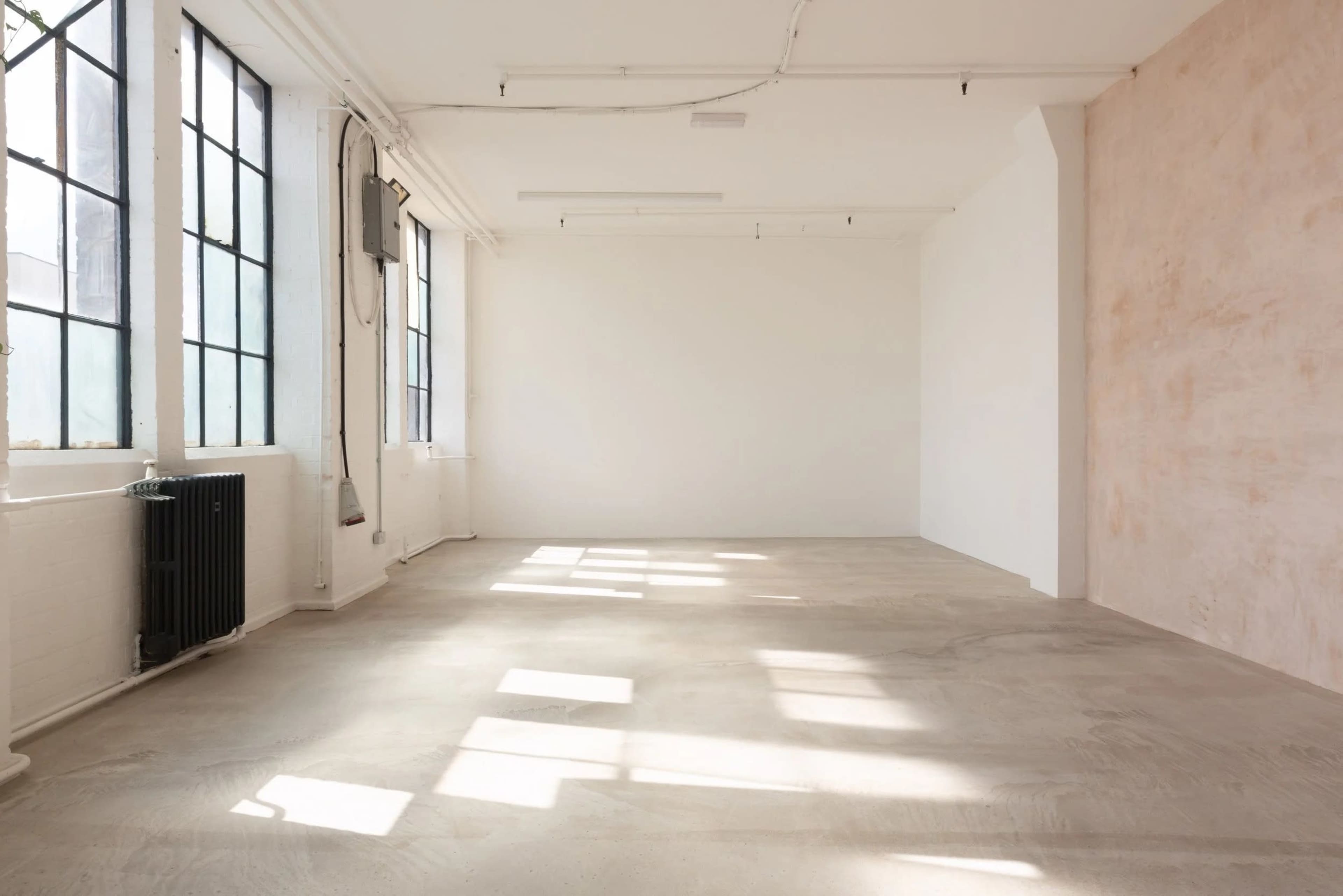 The image shows a spacious, empty room with large windows allowing natural light to illuminate the concrete floor.