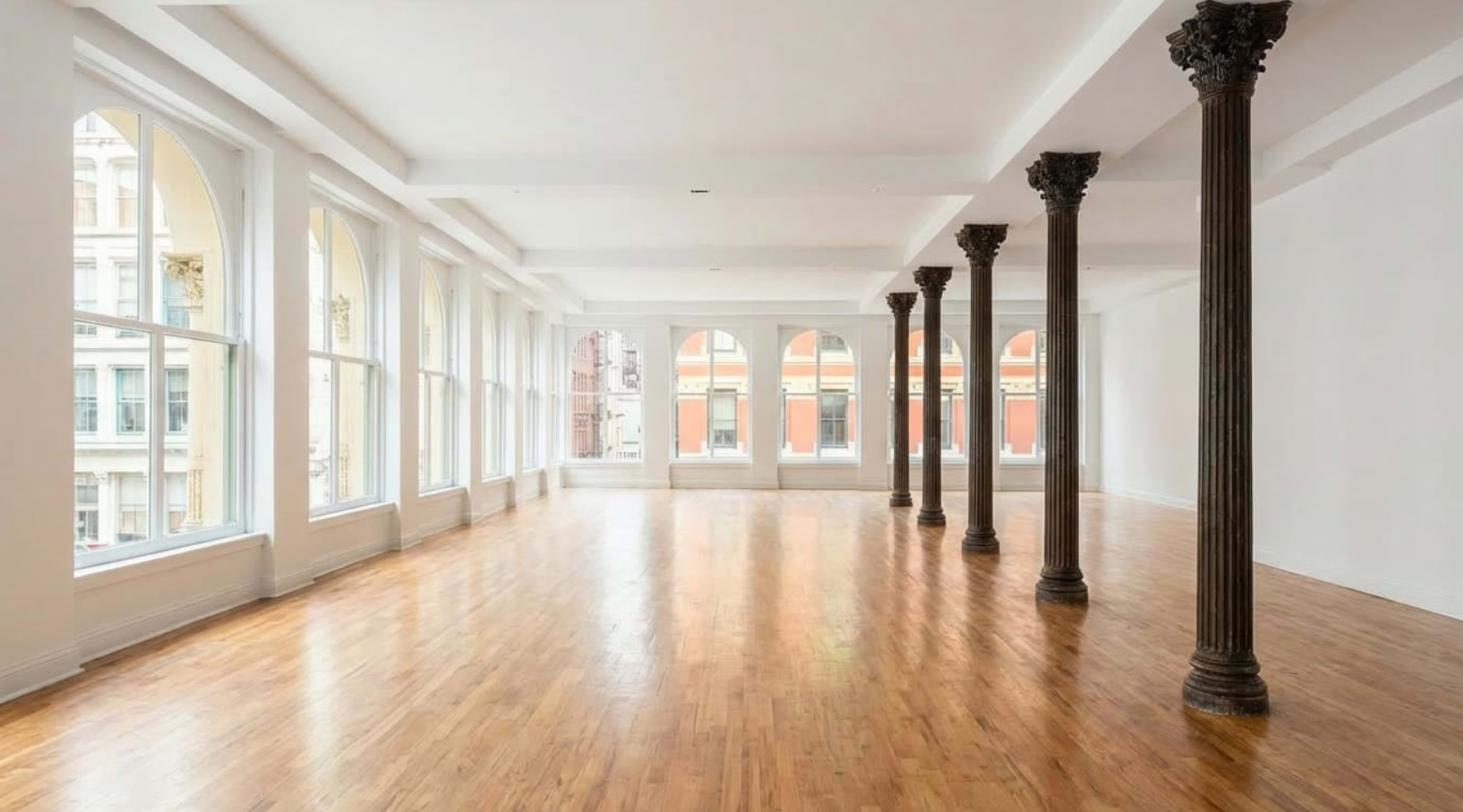 The image shows a spacious, empty room with hardwood floors and large windows, featuring several decorative columns.