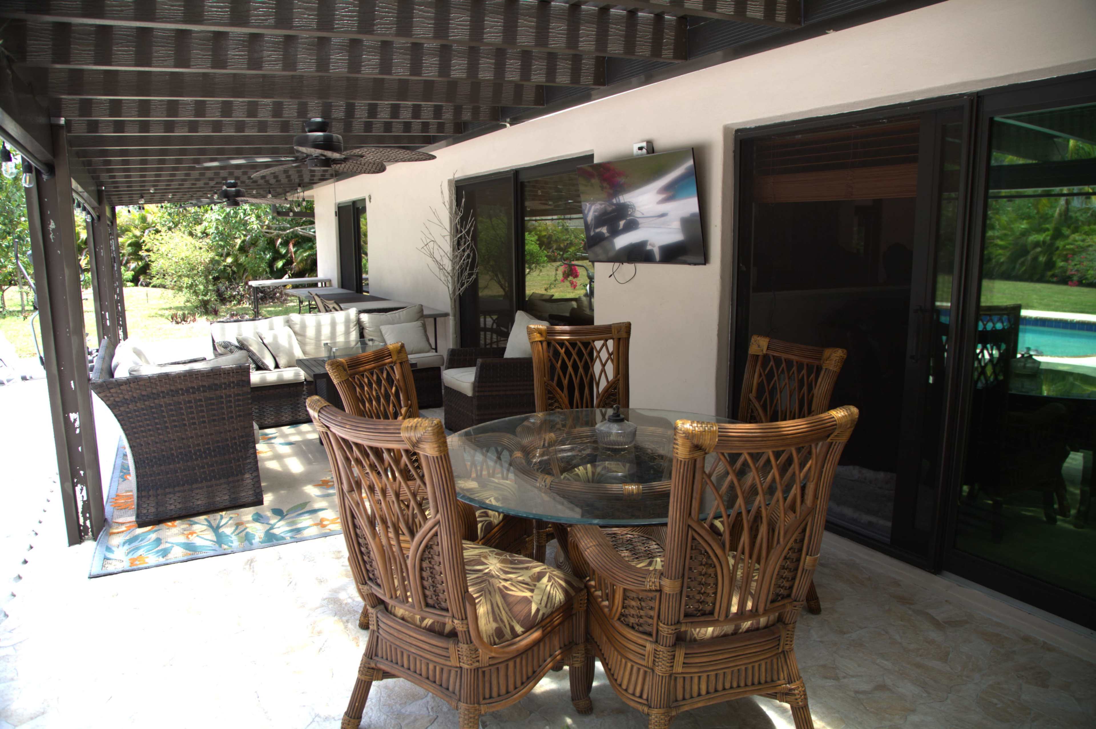 The image shows a patio area with a glass table surrounded by wicker chairs, a lounge seating area in the background, and a television mounted on a wall.
