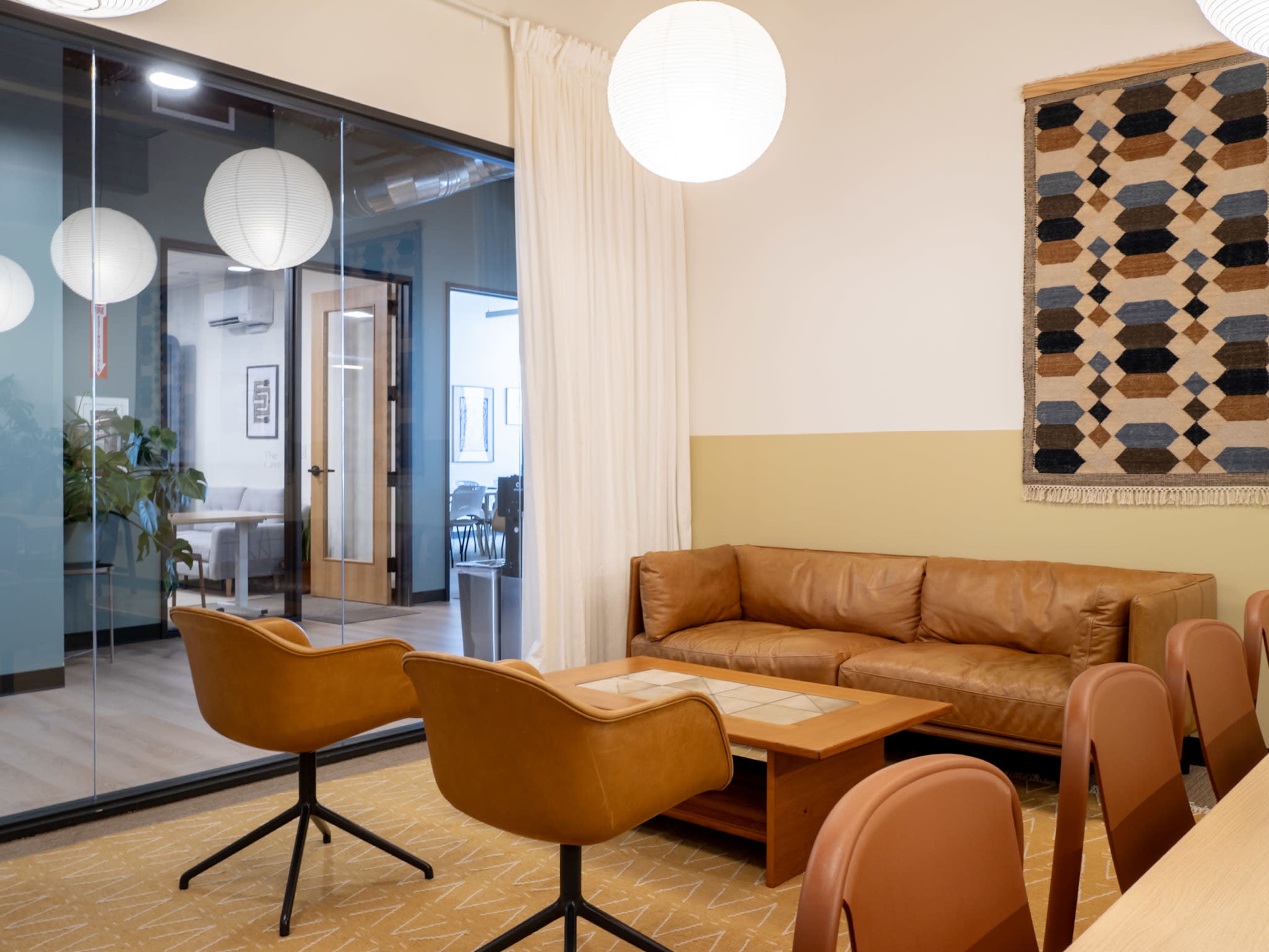 The image shows a modern office space featuring a leather sofa, two mid-century style chairs, a wooden coffee table, and a glass wall separating a meeting room.