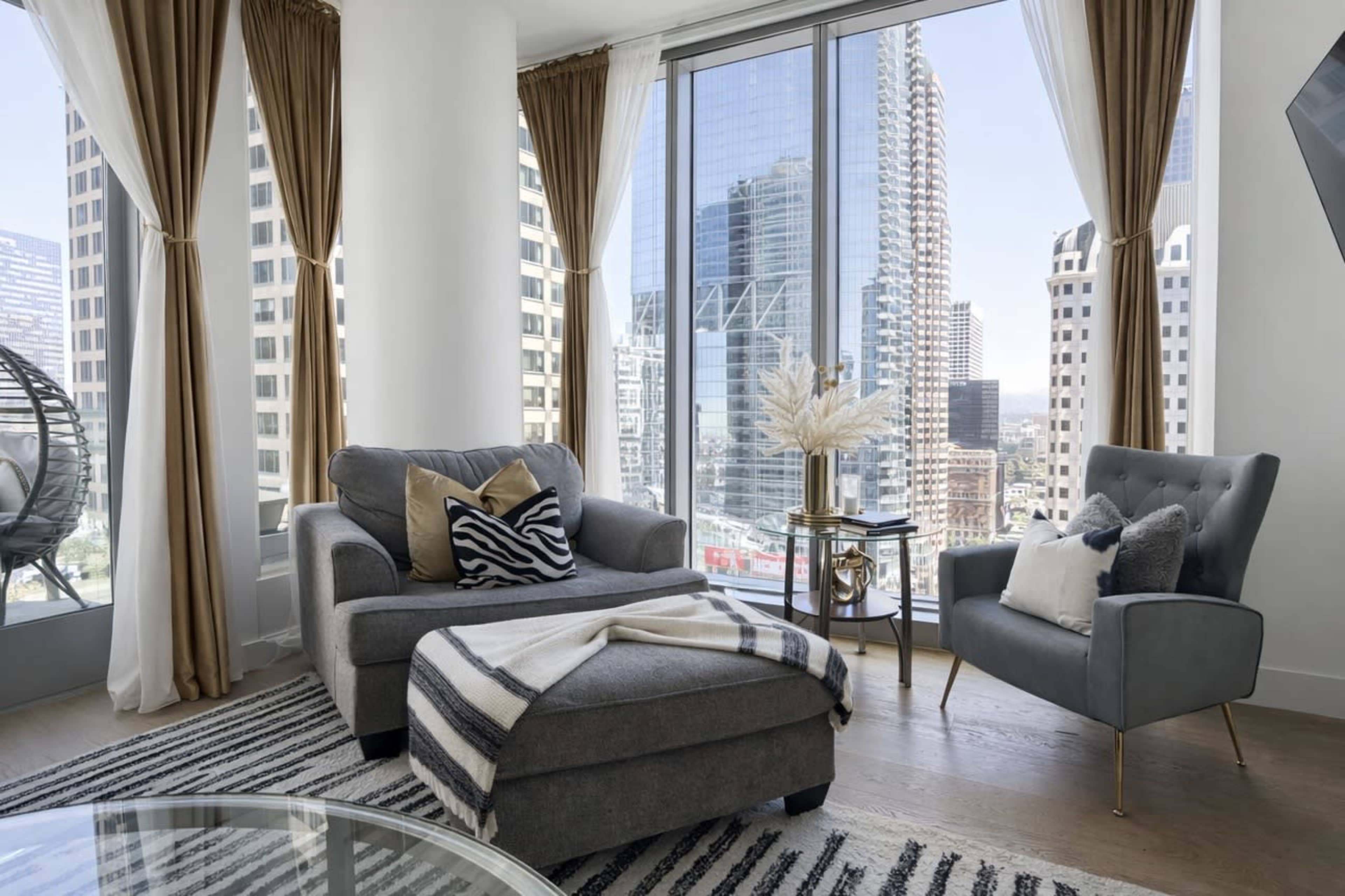The image shows a modern living room with large windows offering a view of city skyscrapers, featuring a gray sofa, an armchair, and a striped area rug.