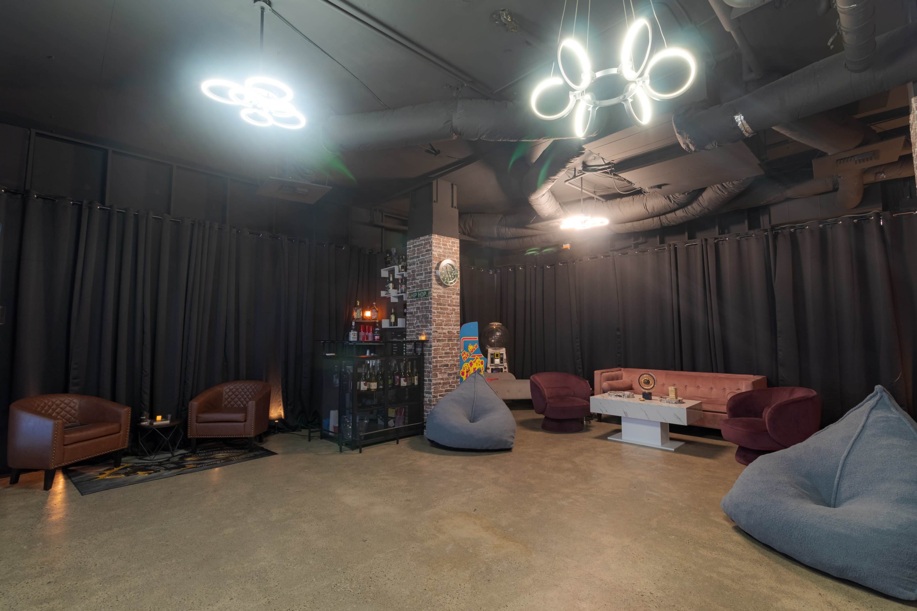 The image shows a modern lounge area featuring bean bags, upholstered chairs, and a minimalist table, surrounded by black curtains and illuminated by contemporary light fixtures.
